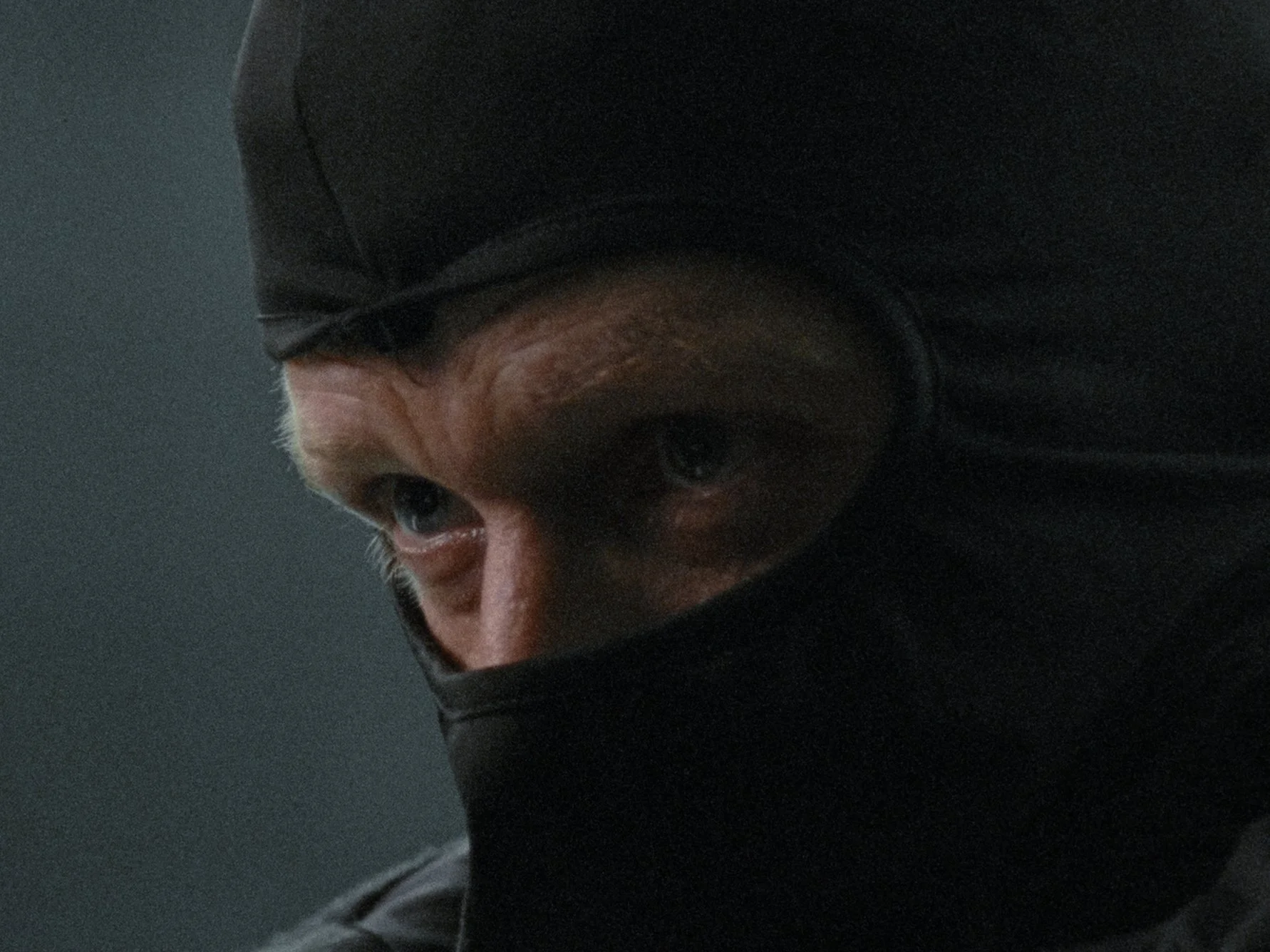 Close-up of a person's eyes and forehead, partially covered with a black face mask and black hoodie, against a dark background.