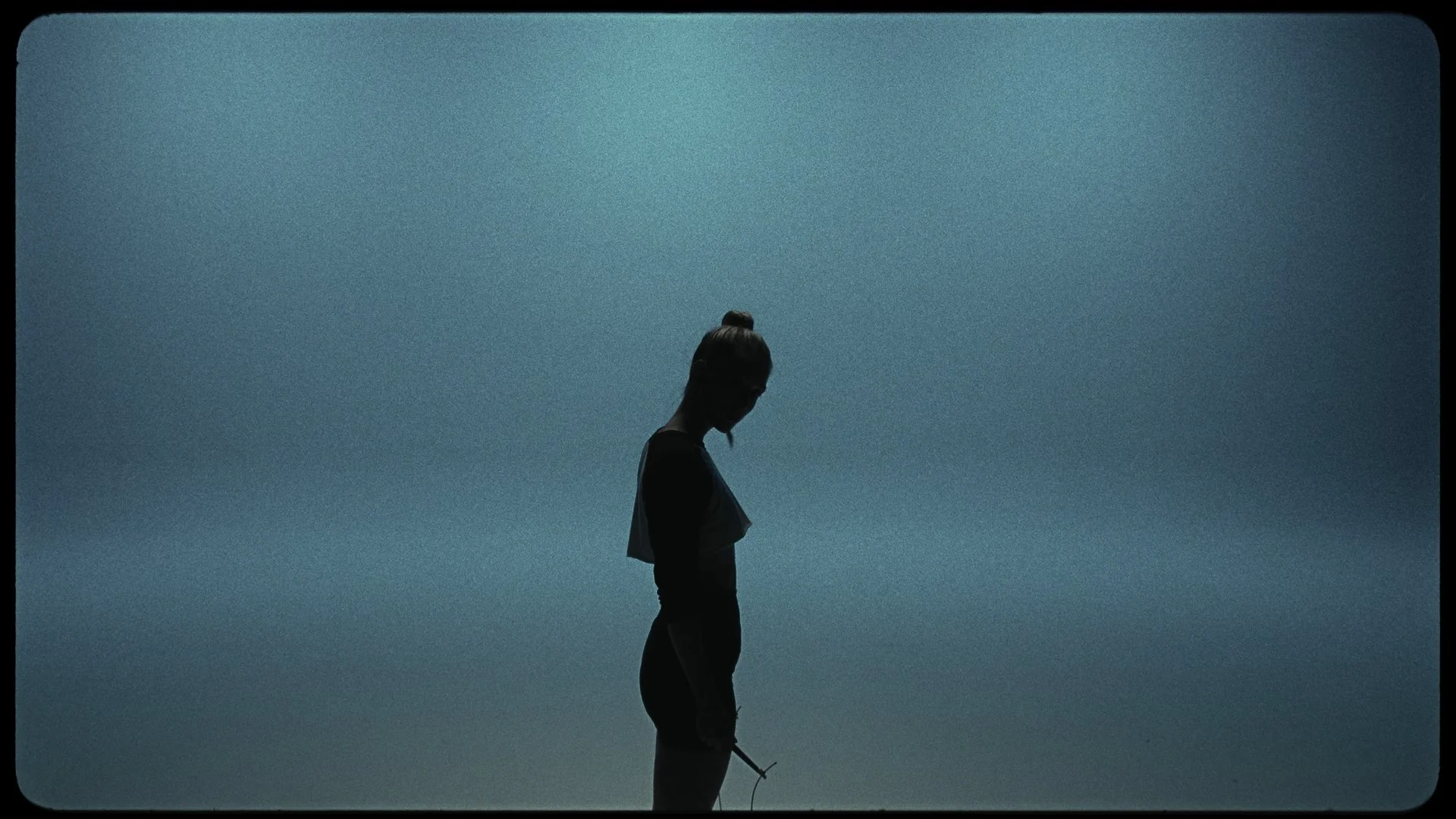 A silhouette of a woman with her hair in a bun standing against a blue sky, looking downward.