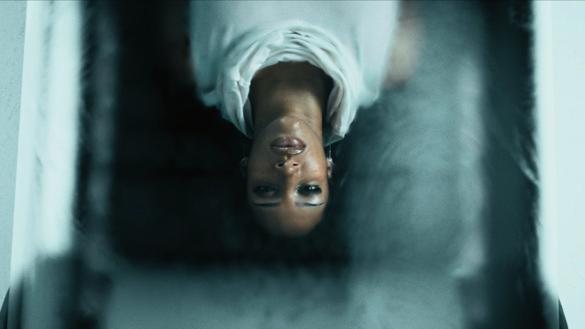 A woman lying on her back, looking up, with her head tilted backwards. She has short hair and is wearing a white top. The photo is taken from her perspective, with her face in focus and the background slightly out of focus.