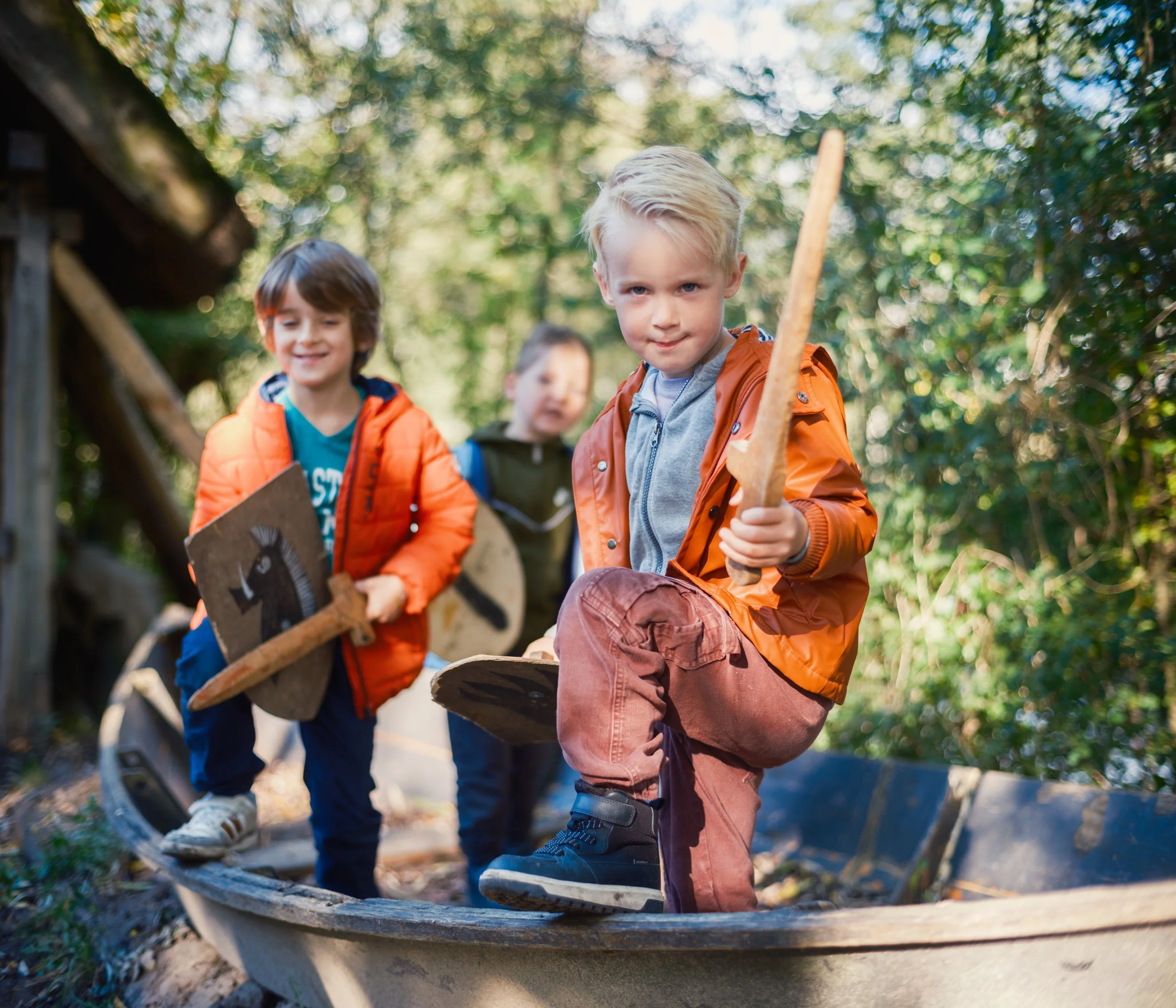 Three children with backpacks playing and walking on a wooden bridge in a forested outdoor setting.