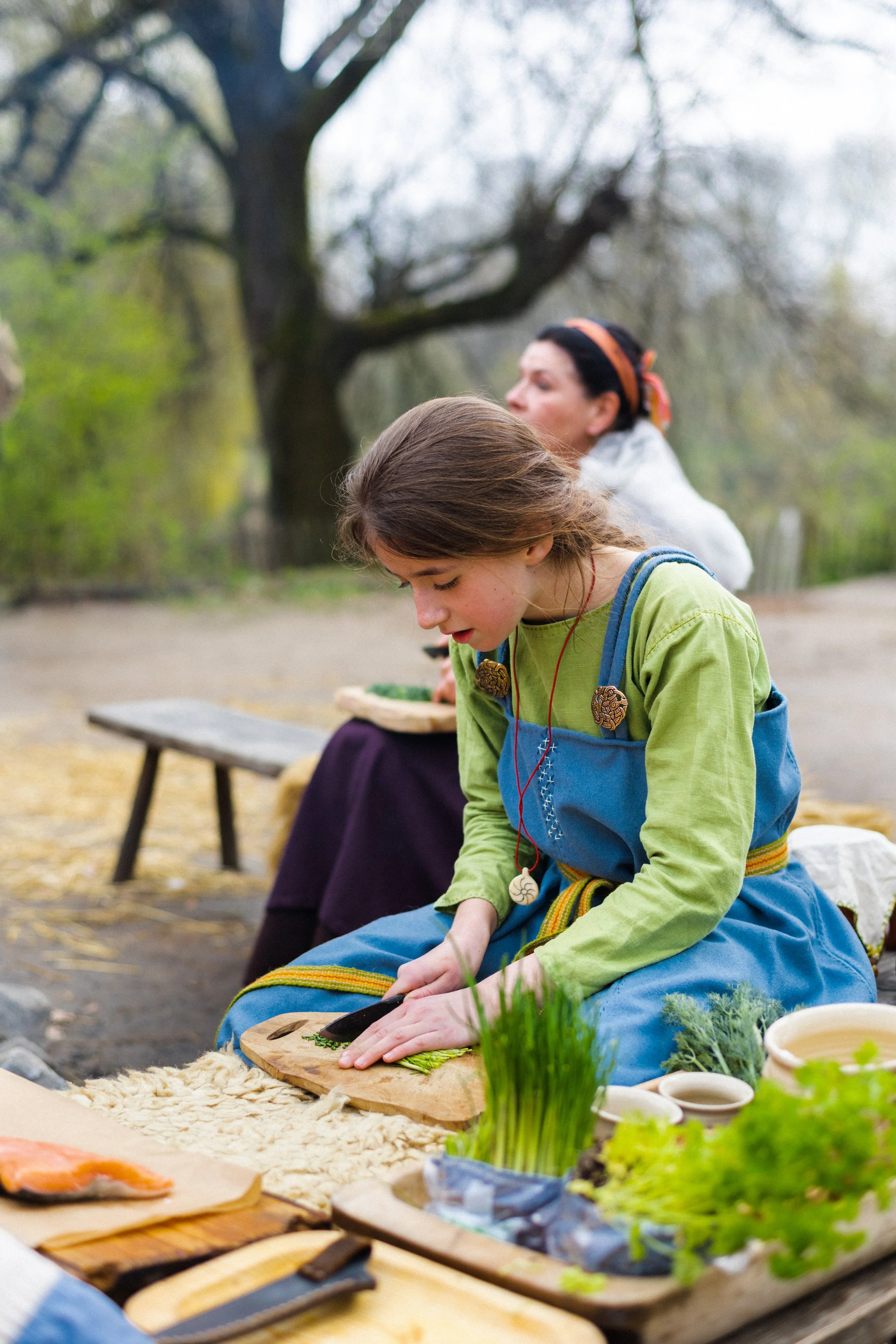 A young girl in a green shirt and blue apron slicing vegetables on a wooden board outdoors during daytime, with a woman in the background also engaged in cooking activities. The setting appears to be a park or garden with trees and natural scenery.