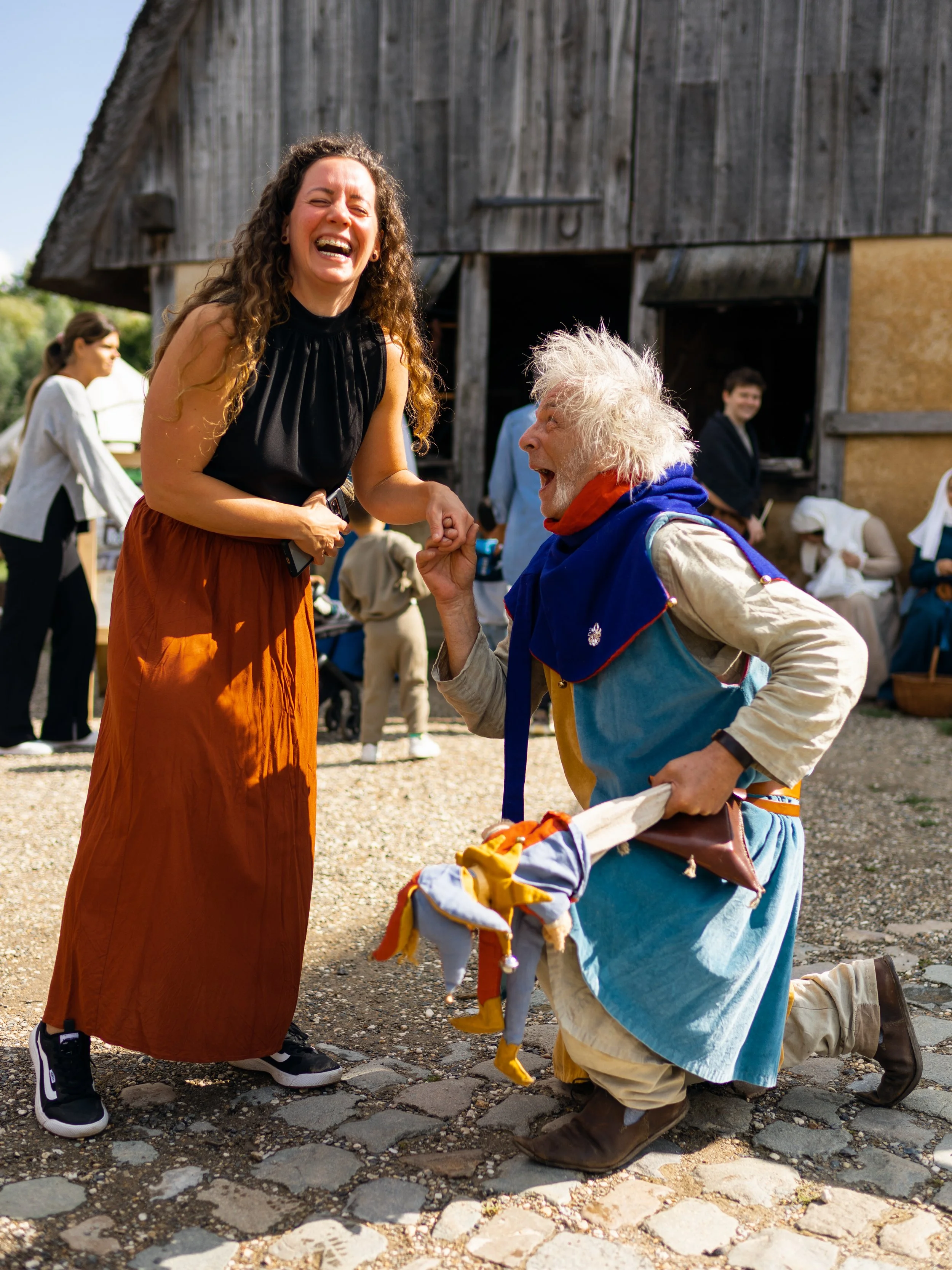 A woman in a black top and orange skirt laughing and holding hands with an older man who is kneeling and dressed in colorful, layered clothing, outdoors near a barn with other people in the background.