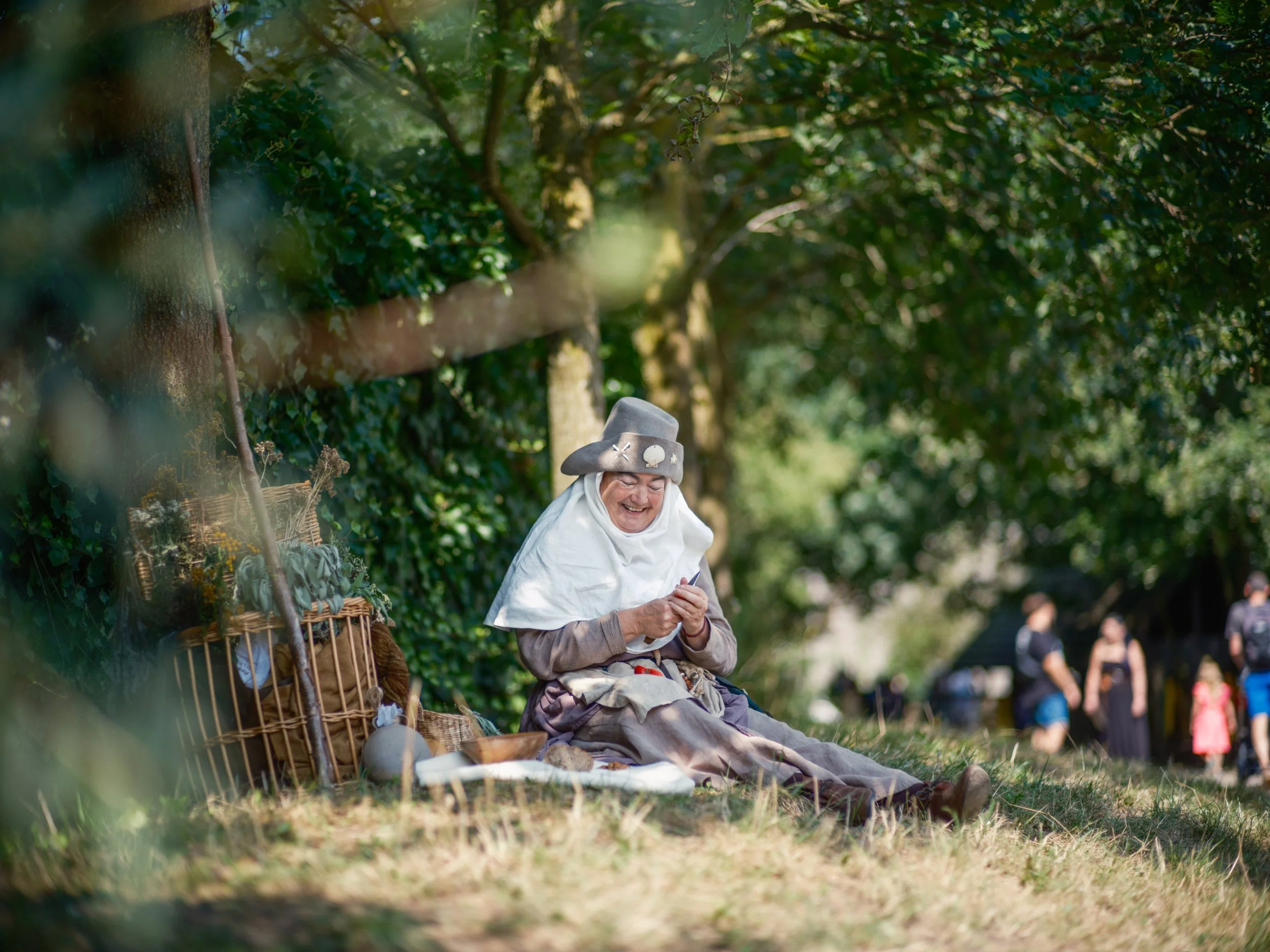 An elderly woman dressed in period or historical attire, sitting on the grass in a park, smiling while looking at her phone. She is wearing a wide-brimmed hat decorated with patches and a loose, layered skirt. There are baskets and historical objects