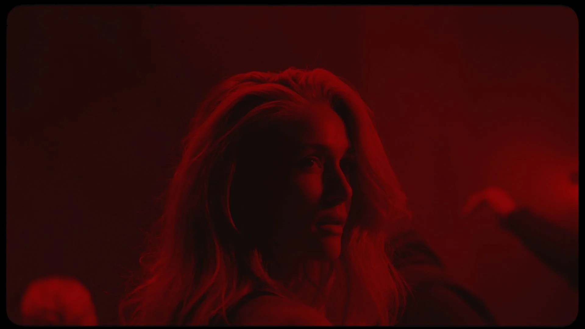 Woman with blonde hair illuminated in red lighting, looking to the side with a serious expression.
