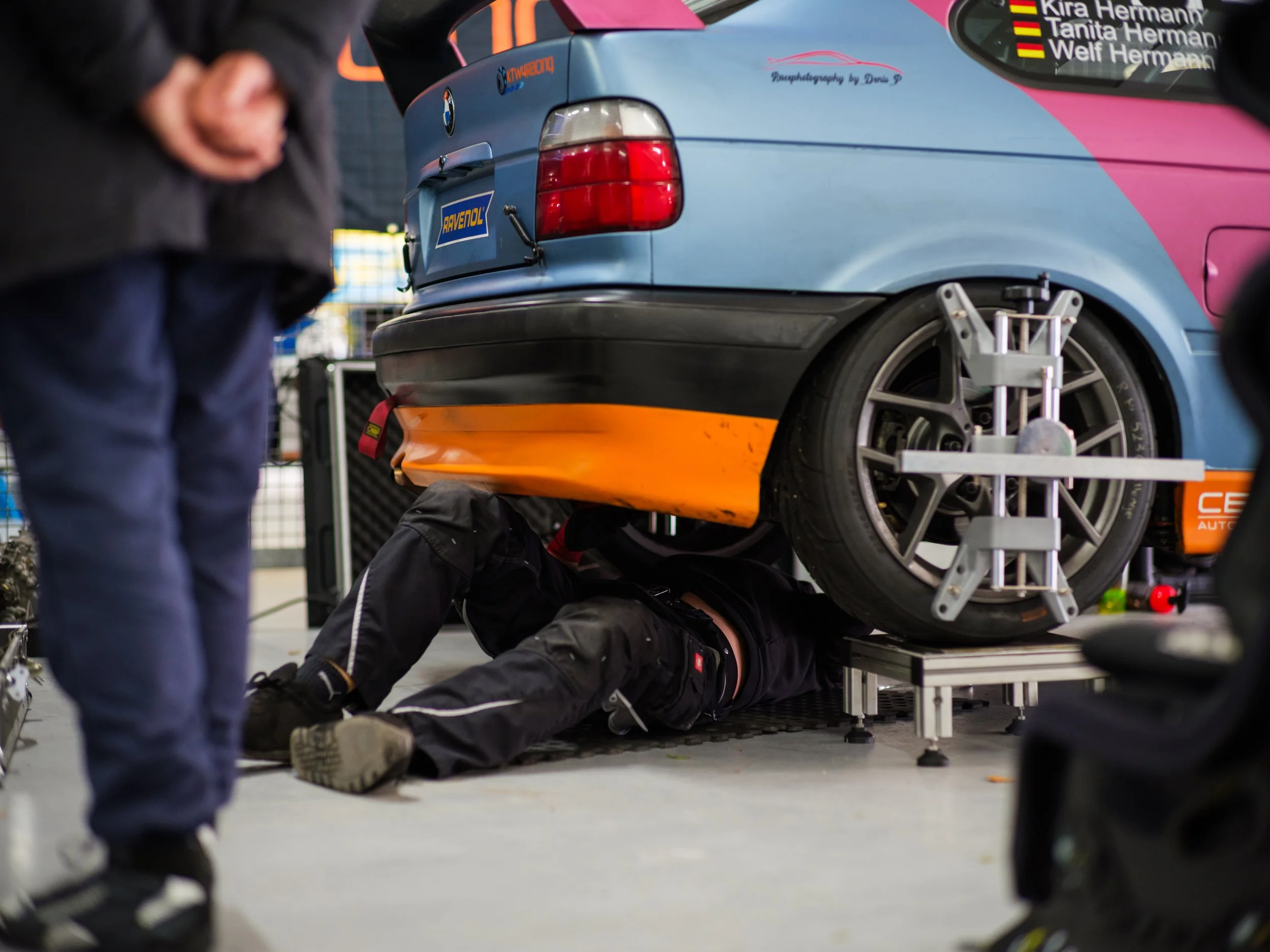 A race car on a lift with a mechanic working underneath, with another mechanic in the foreground.