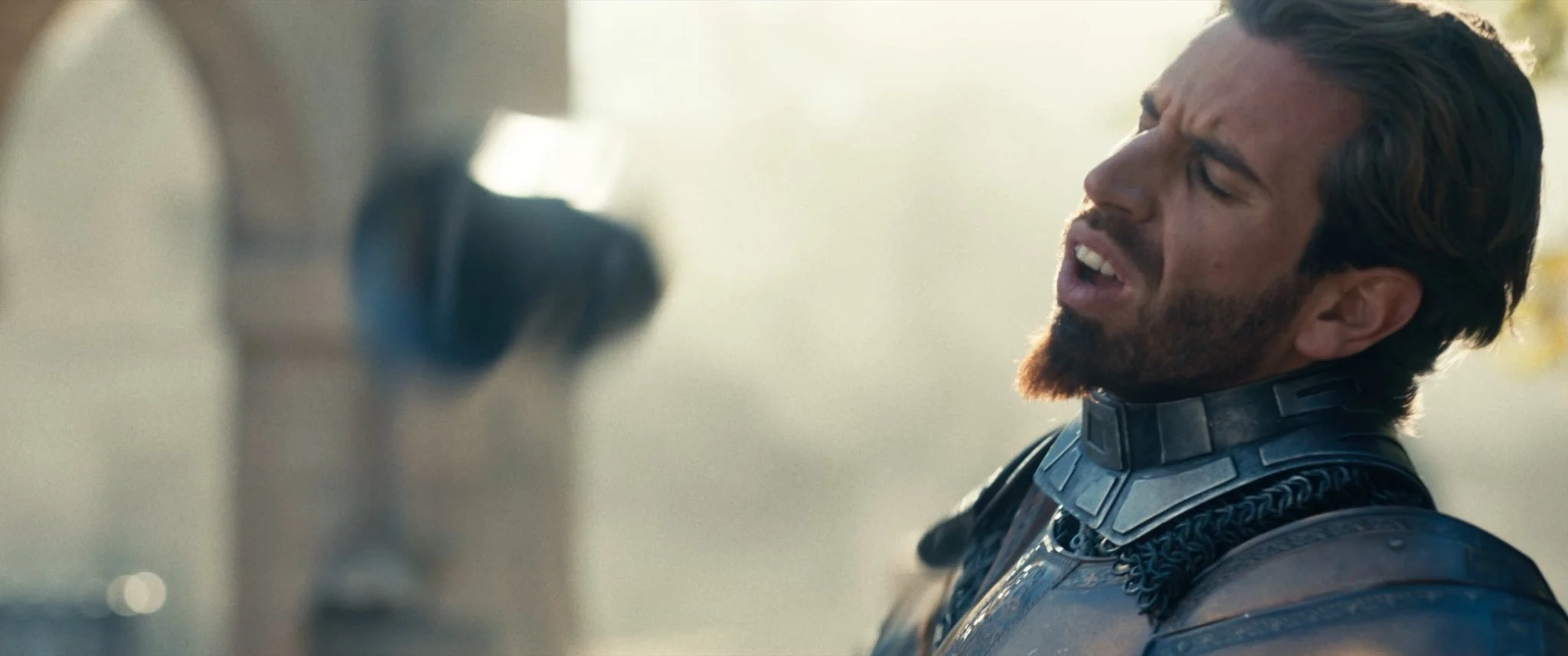 Close-up of a man with dark hair and beard wearing a futuristic armored suit, appearing distressed or in pain, with blurry background.
