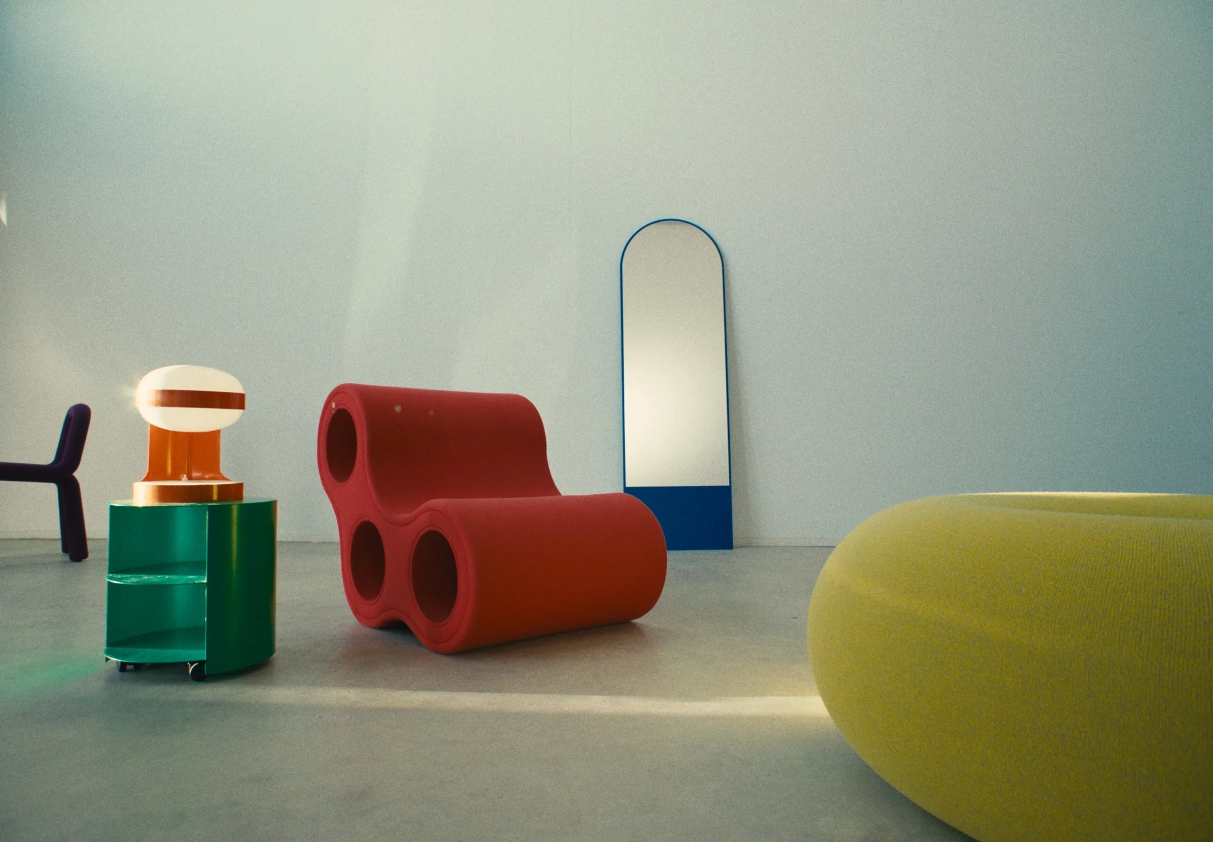 Minimalist interior with colorful modern furniture including a yellow chair, green and black storage units, a red bench with circular cutouts, a rounded white and orange lamp, and arches with a mirror, set against a plain light-colored wall.