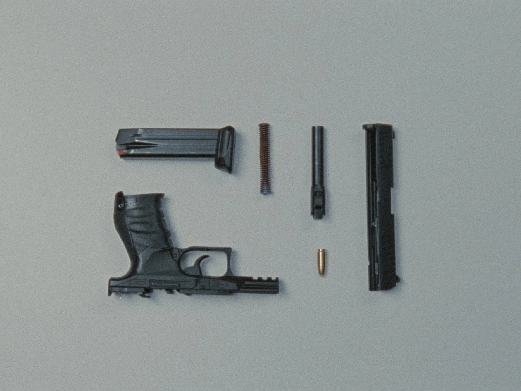 Disassembled handgun with magazine, spring, firing pin, and other firearm parts laid out on a light gray background.