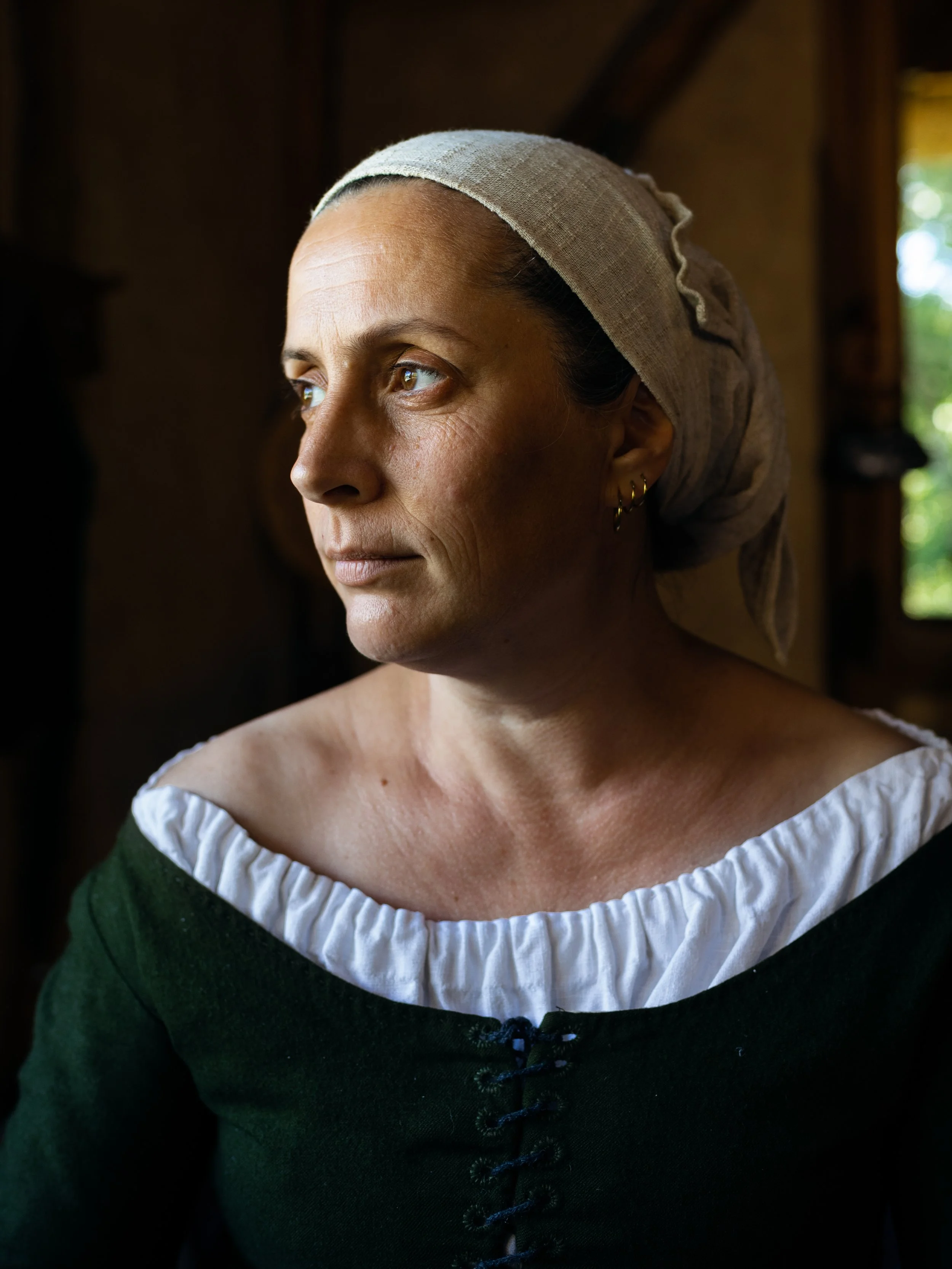 Close-up of a woman with light skin, wearing a beige bonnet and a black dress with a white ruffled collar, looking to her left inside a room with wooden beams and natural light.