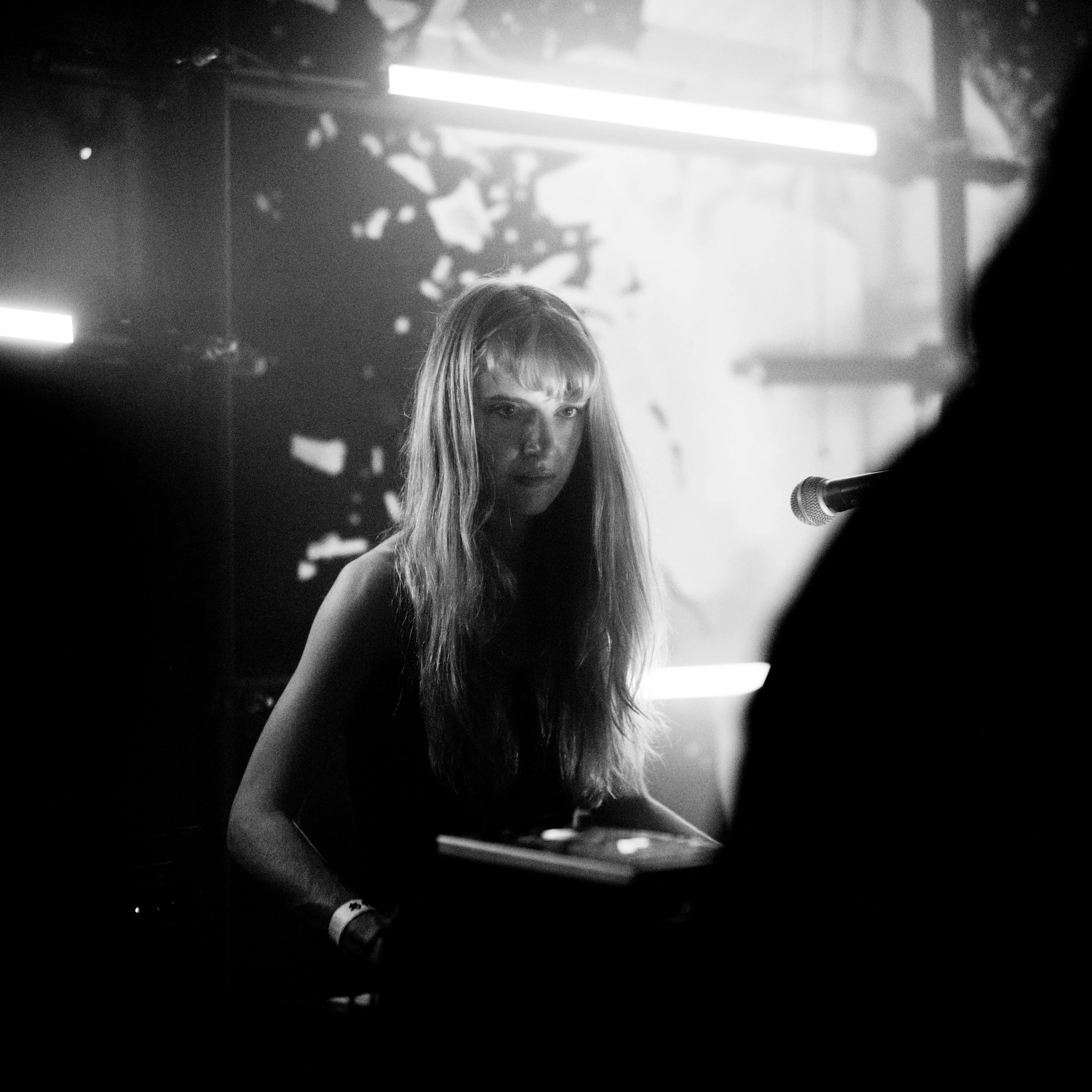 Black and white photo of a woman with long hair, looking at the camera, with a microphone in front of her, indoors with light sources and shadows creating a moody atmosphere.