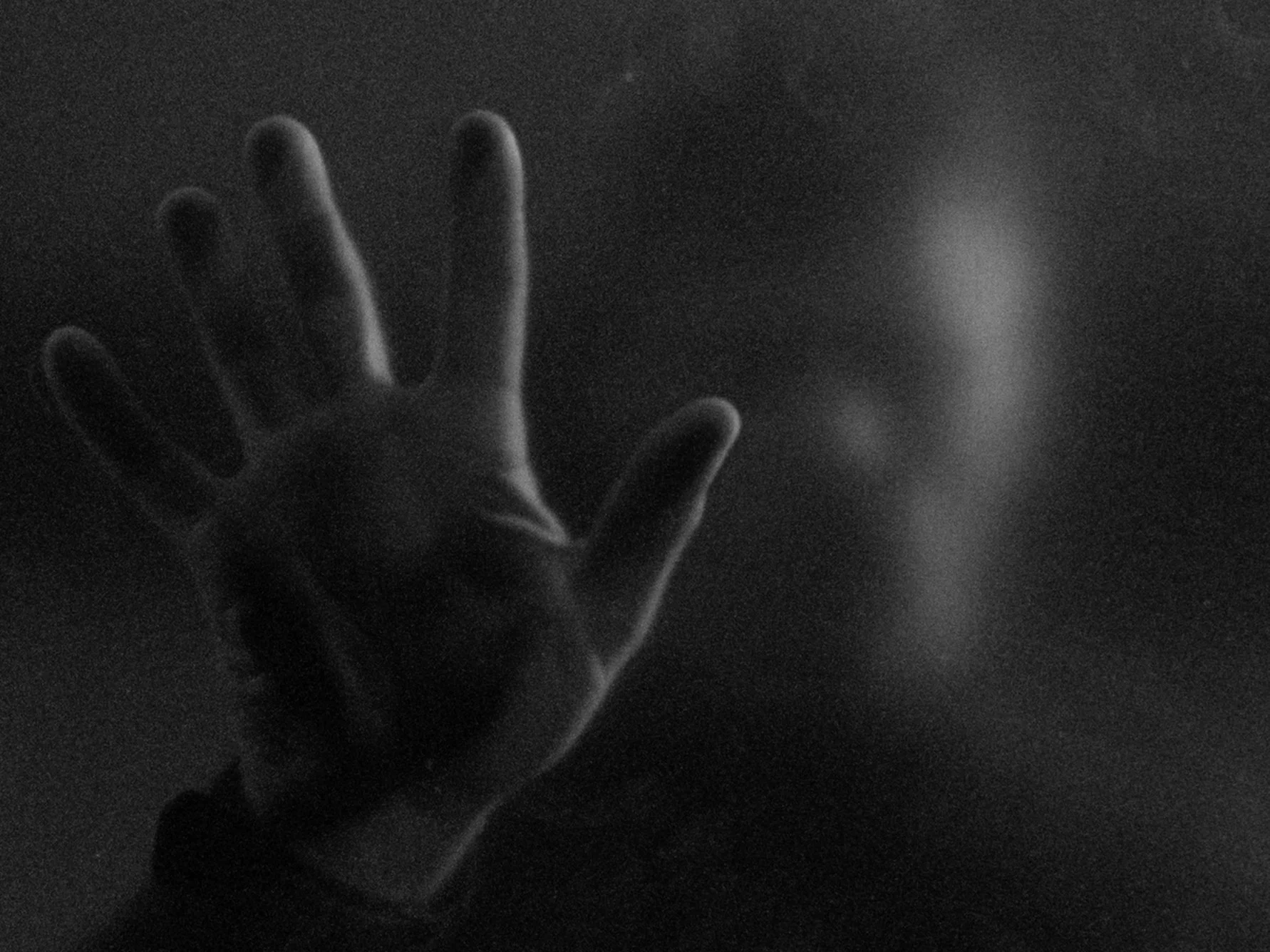A black and white photograph of a hand with fingers slightly spread, reaching upwards, against a dark background with soft lighting.
