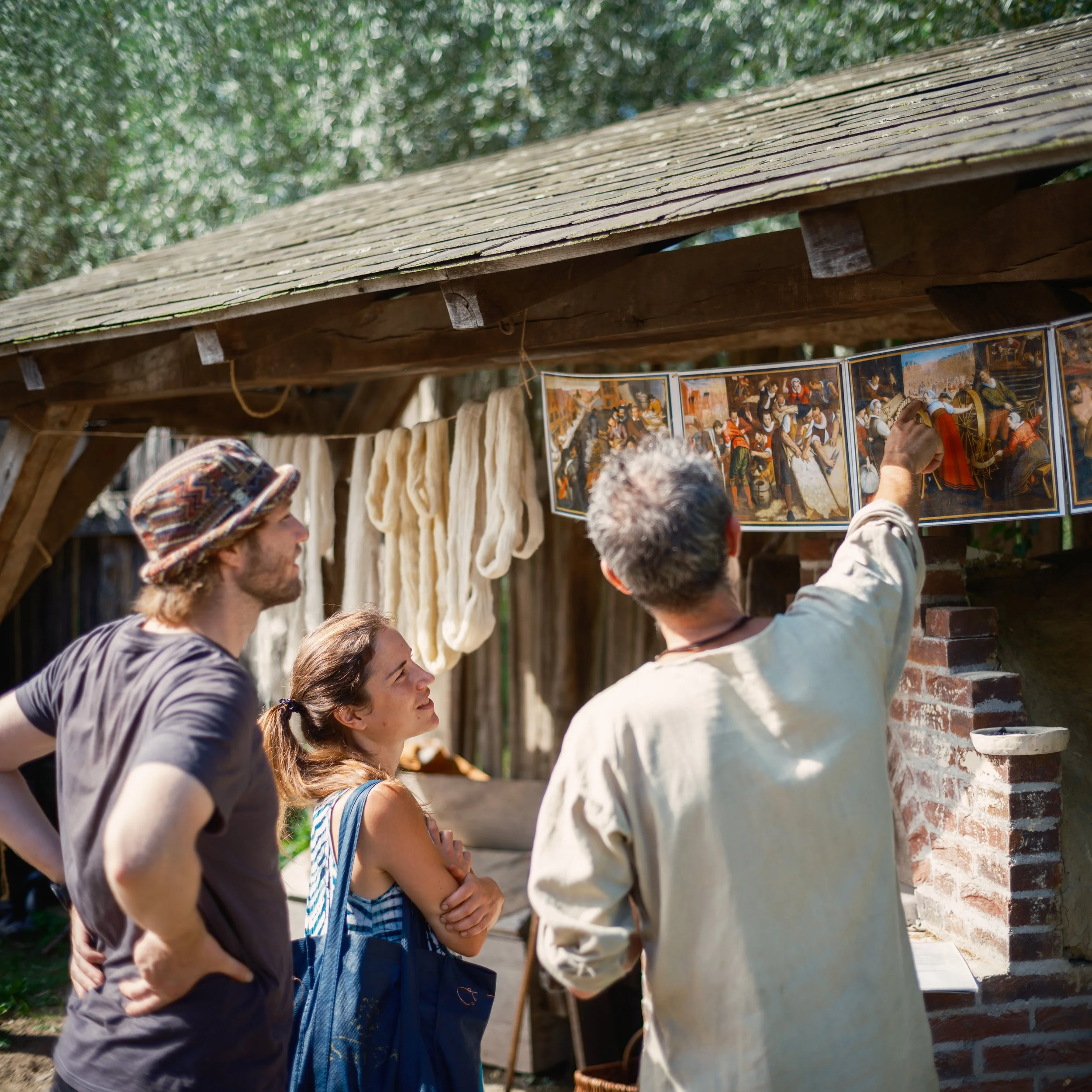Group of people gathered outdoors, observing a person showing something on a wall with artwork images, under a wooden shelter with hanging cloths and greenery in the background.