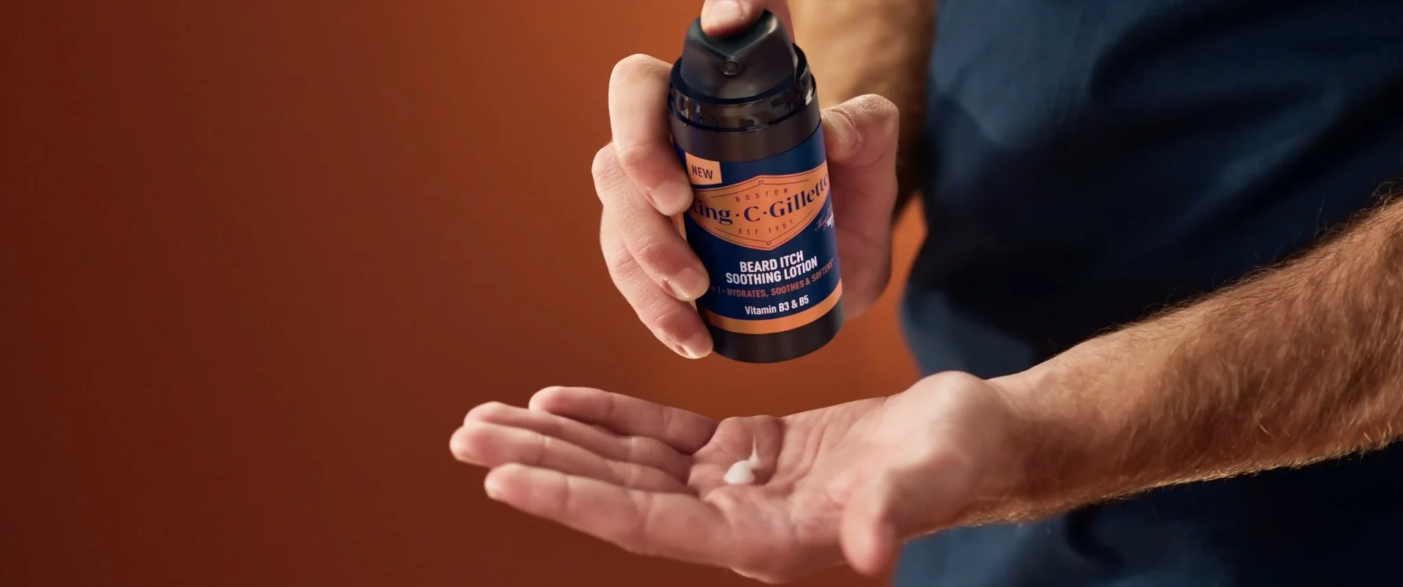 A person dispensing lotion from a dark bottle labeled 'King C. Gillette Beard Itch Soothing Lotion' onto their hand, with a brownish background.