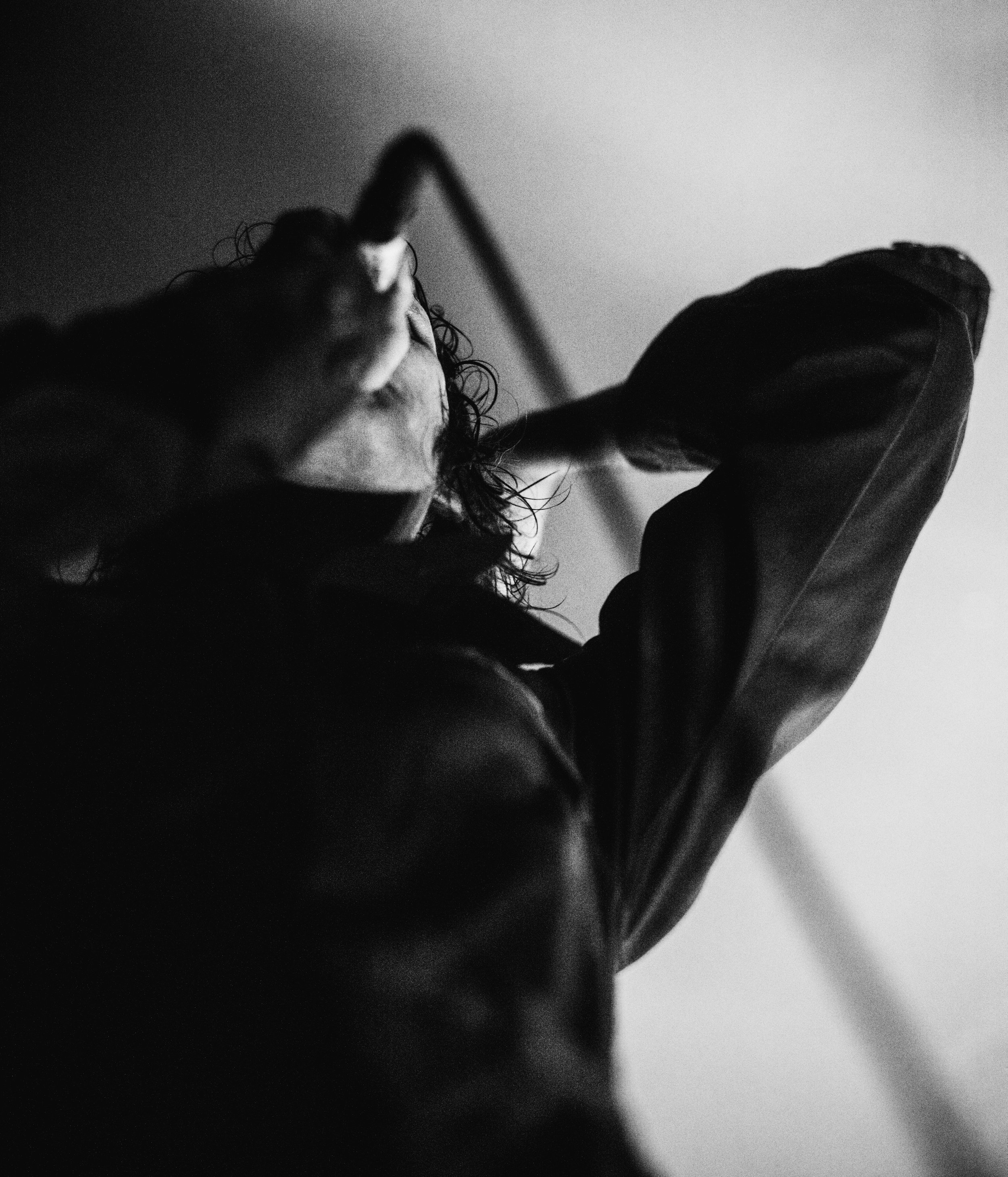 Black and white photo of a person taking a selfie from below, with curly hair, wearing a jacket, with a plain background.