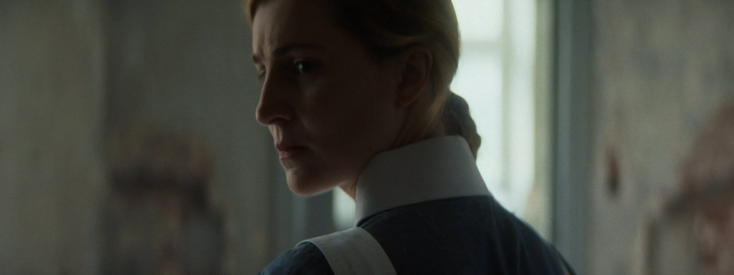 A woman with red hair, dressed as a nurse, looks over her shoulder in a dimly lit room.