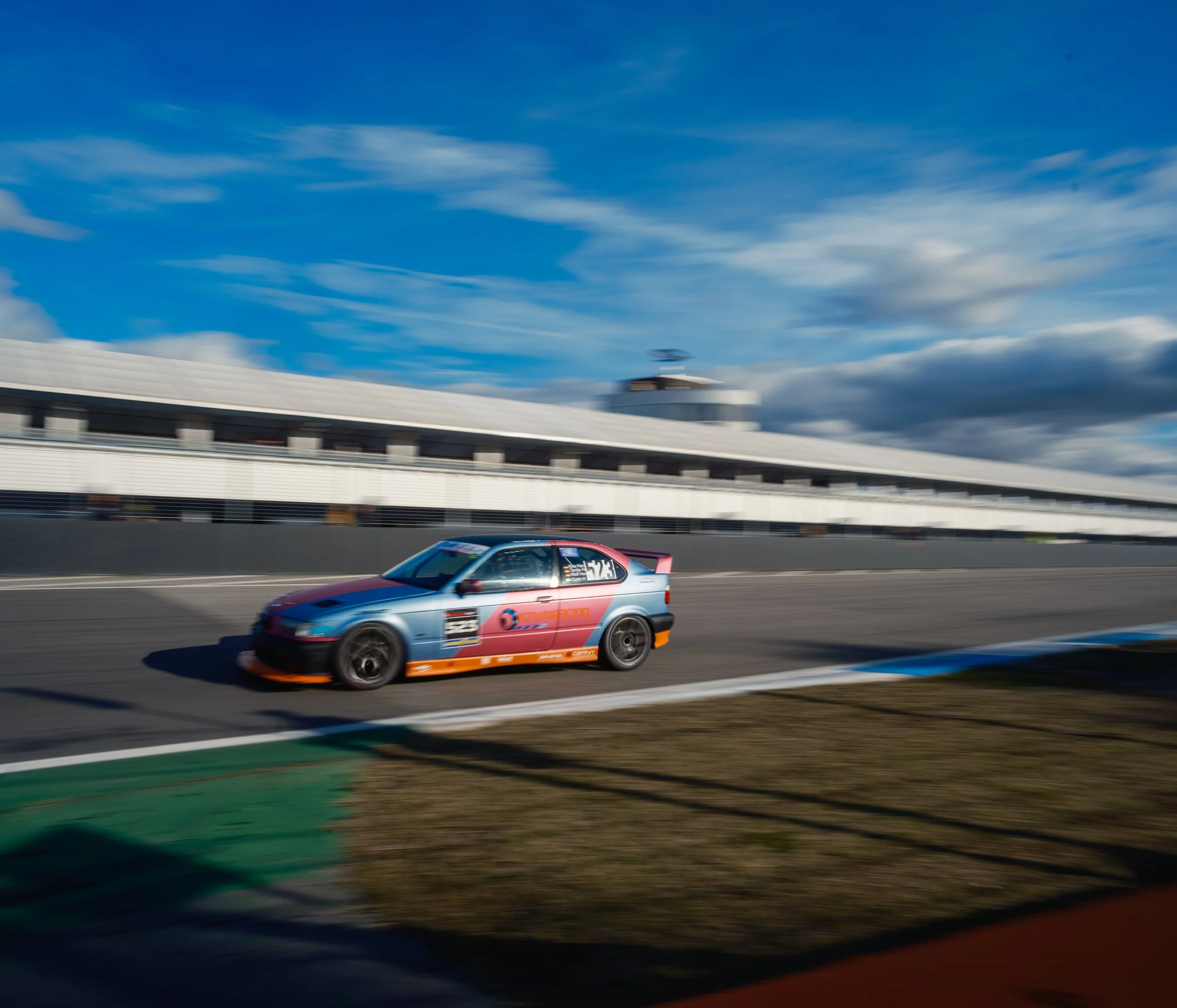 A race car speeding on a racetrack with a blurred background of a modern stadium and sky with scattered clouds.