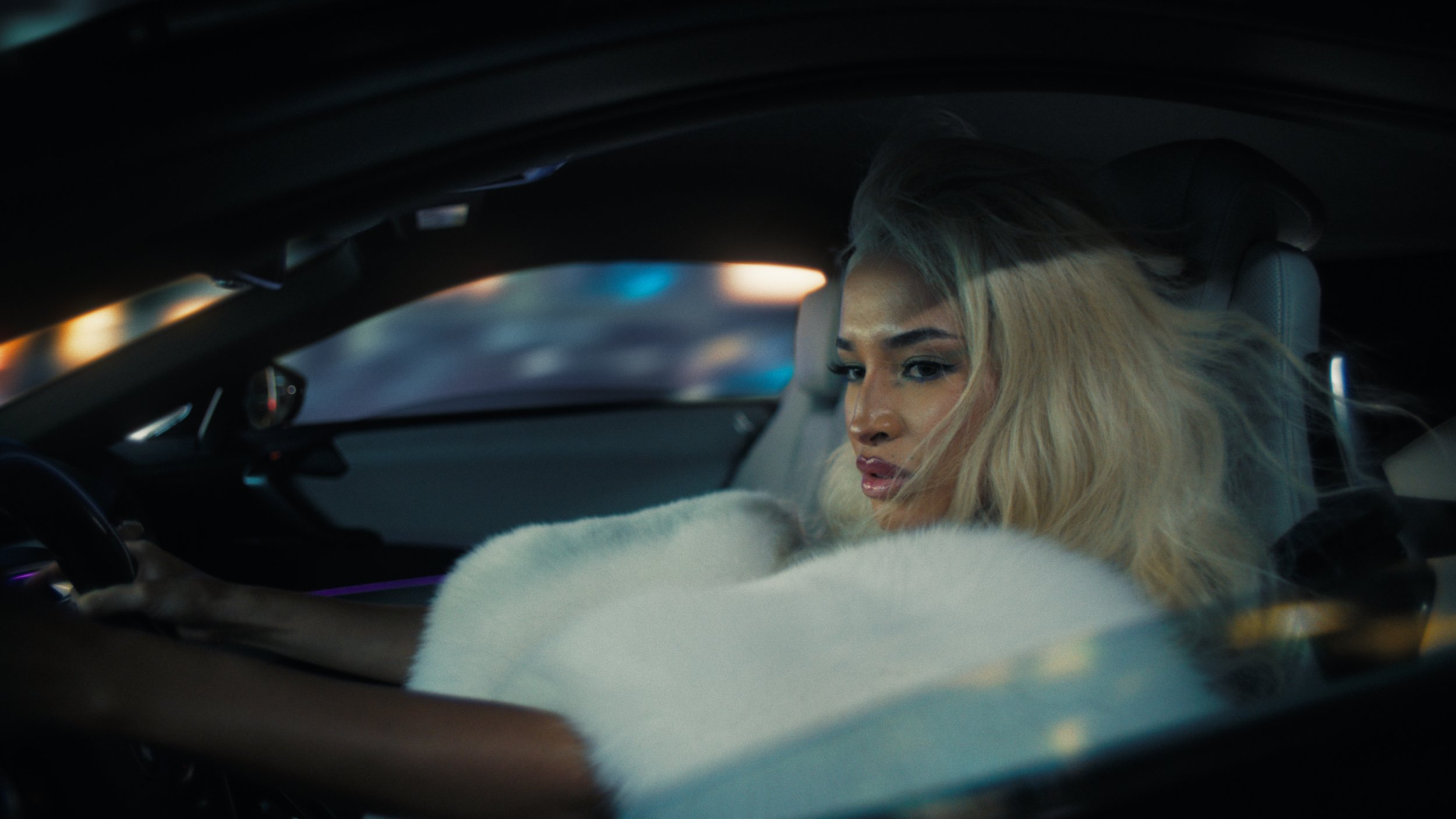 A woman with blonde hair and makeup inside a car, looking out while resting her arm on the window