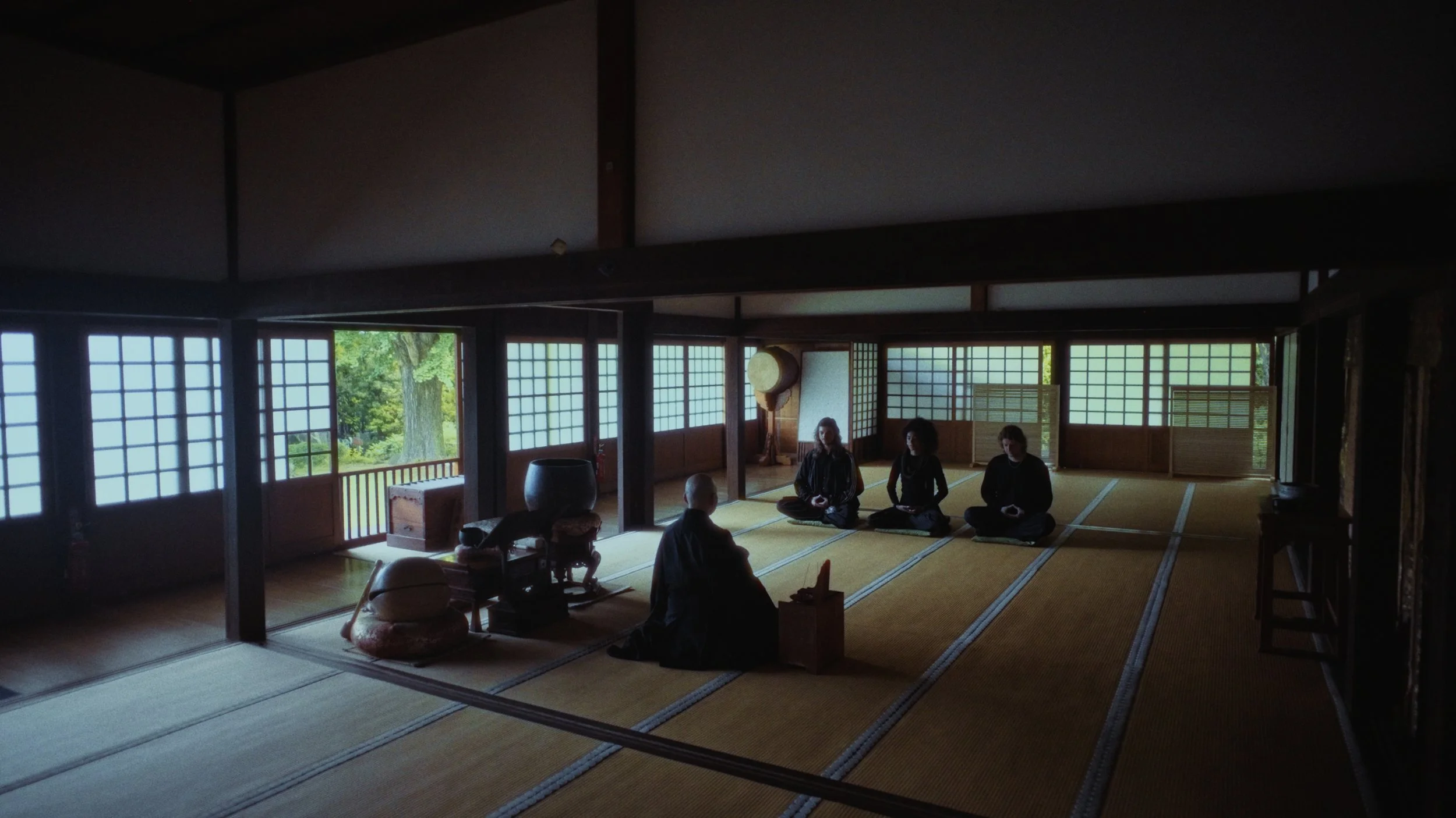 Interior of a traditional Japanese room with tatami mats, shoji screens, and three people seated in a meditative posture. Outside, green trees are visible through the open windows.
