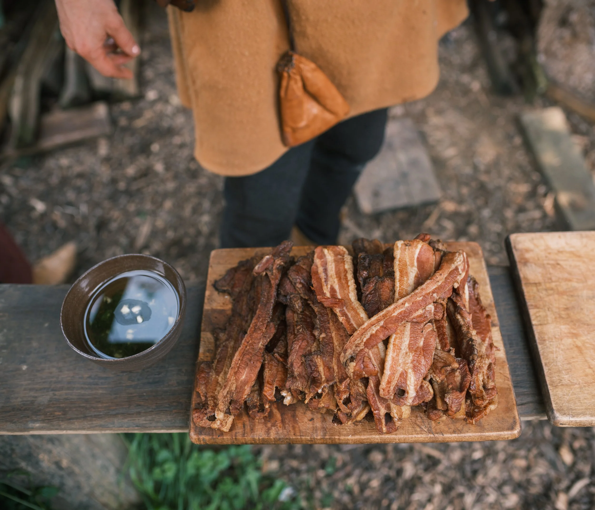 A wooden board with sliced cooked bacon on it, next to a glass of dark beverage with ice and a lime wedge, outdoors on a rustic table.