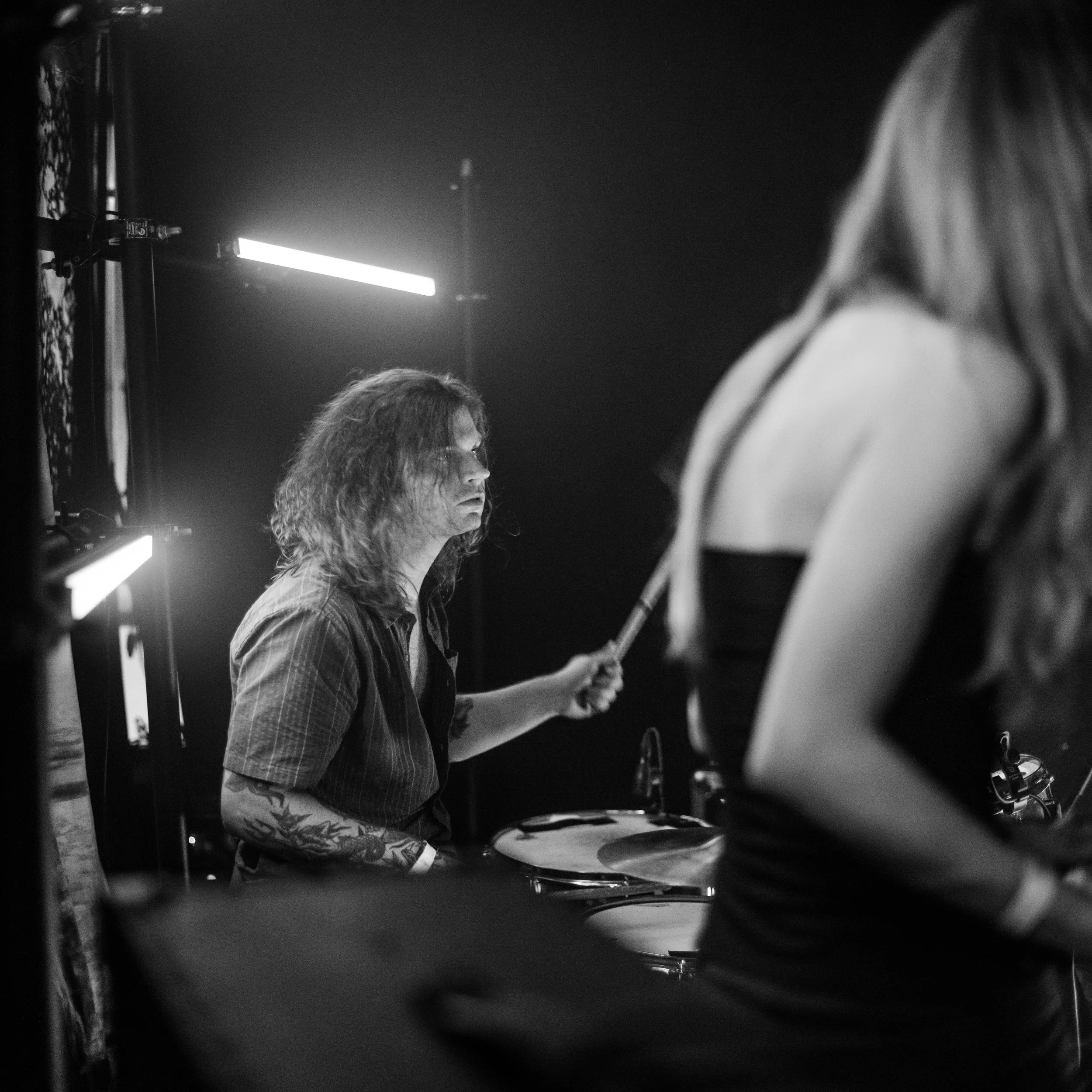 Black-and-white photo of people, including a woman with wavy hair, in a dark indoor setting with bright lighting. The image appears to be taken through a reflective or transparent surface.