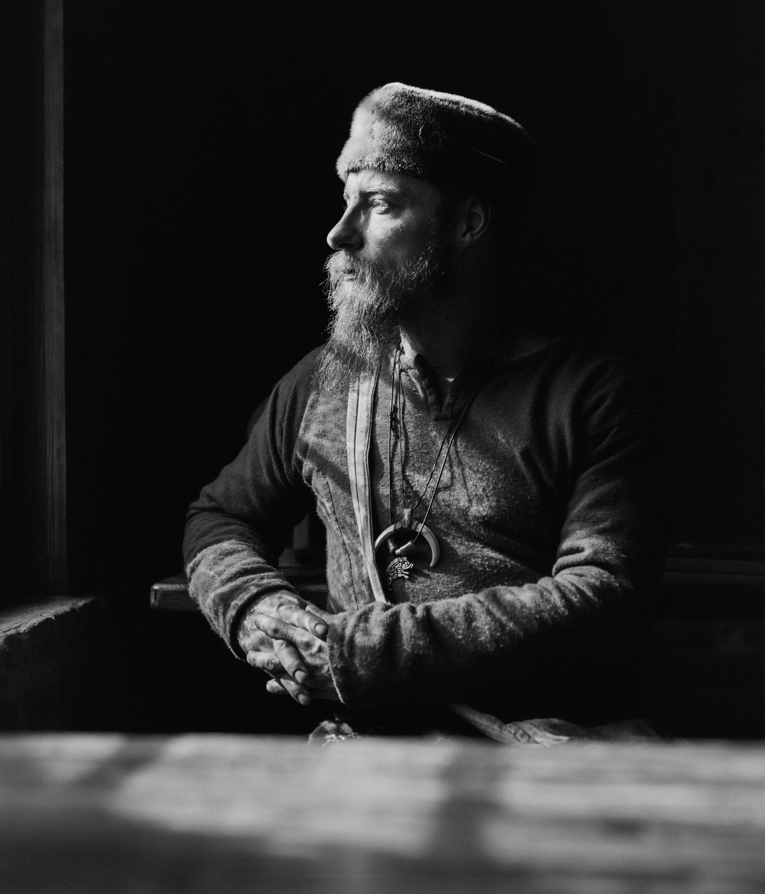 Black and white photo of a man with a beard wearing a fur hat, looking to the right, with a serious expression. The background is dark, and he is illuminated by a soft light from the left side.