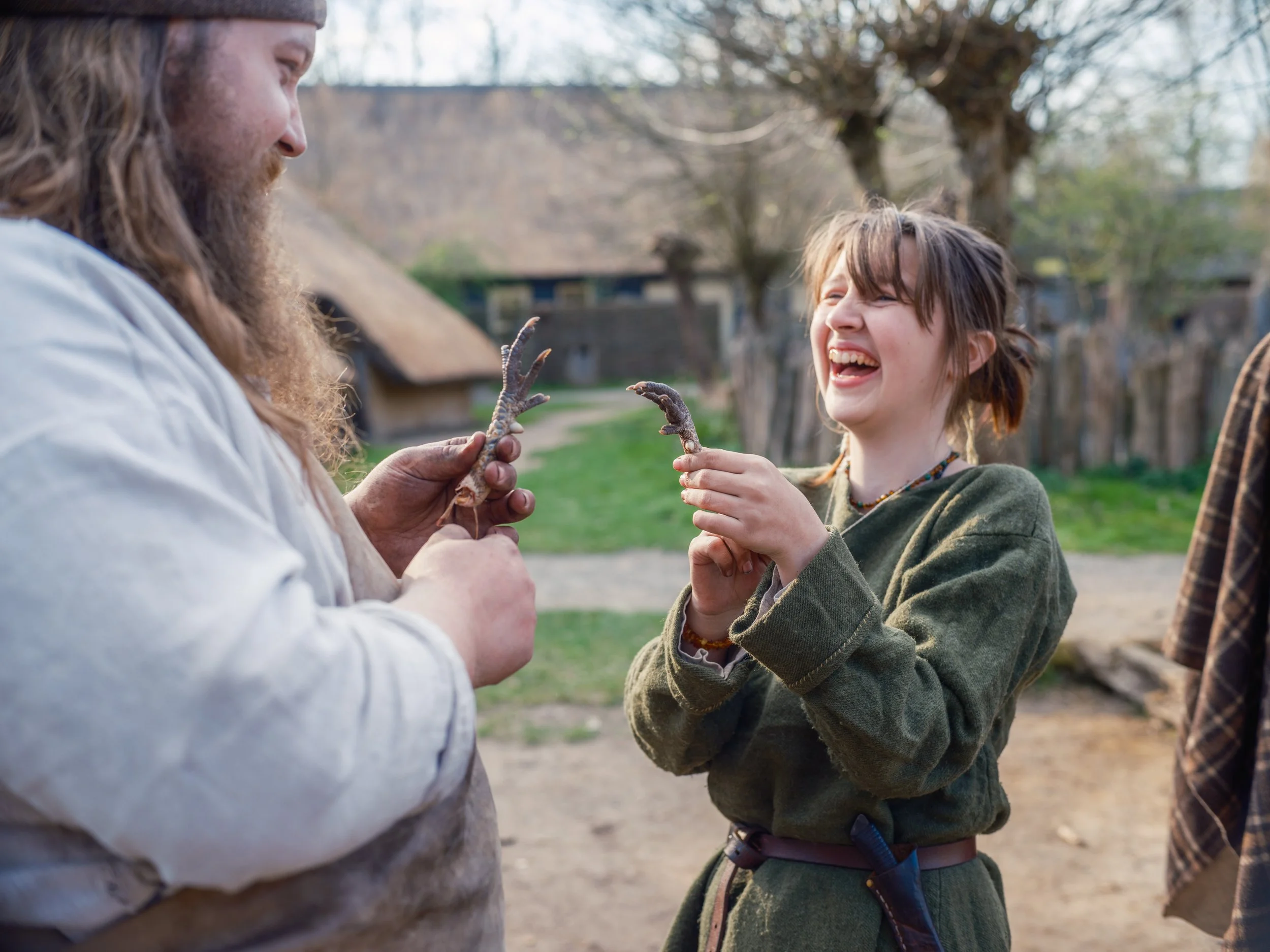 A man with long hair and a beard holding a small animal, possibly a lizard or an amphibian, while a young girl with short hair and a green jacket interacts with it outdoors in a natural setting with trees and houses in the background.