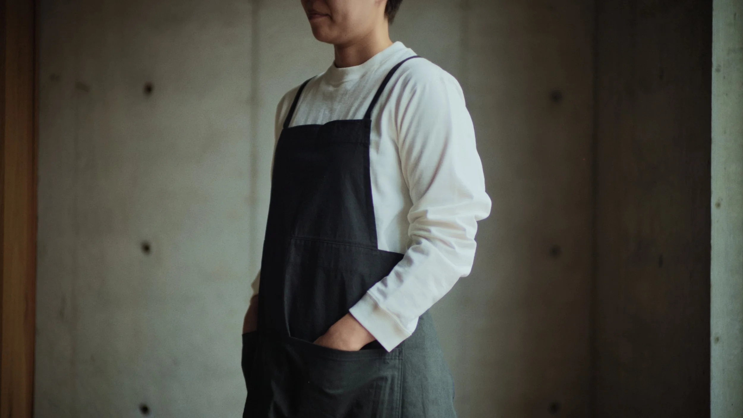 A person is standing against a concrete wall wearing a white long-sleeve shirt and a black apron, with one hand in their pocket and a slight smile.
