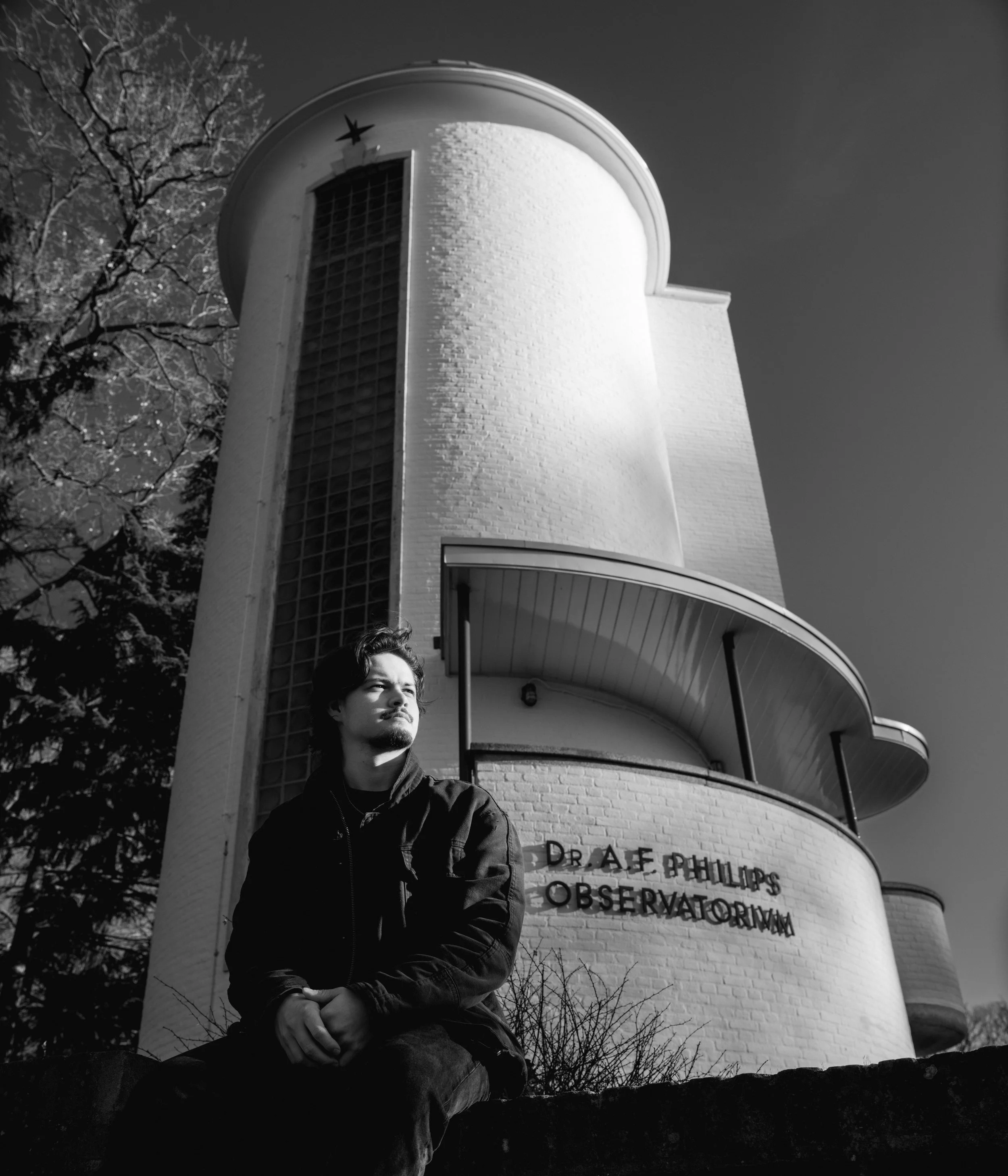 A young man with dark hair and a beard, wearing a dark jacket, sitting on a ledge outside a white brick building with a cylindrical tower and a sign that reads 'DR. A.F. PHILLIPS OBSERVATORY.' Trees without leaves are visible, and the sky is clear.