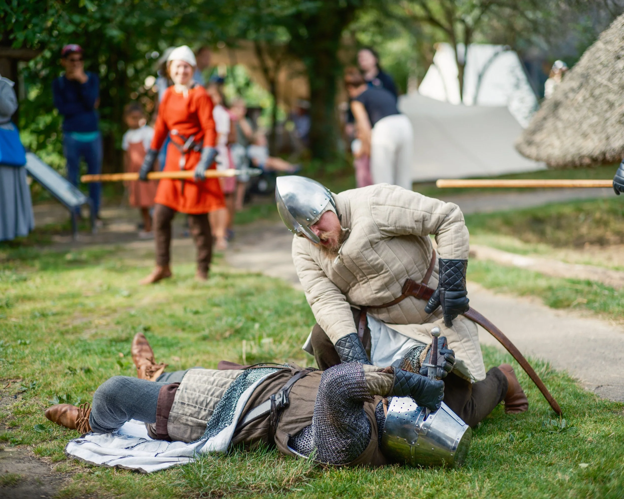 A reenactment of a historical battle with people dressed in medieval armor, some lying on the ground and others standing behind them, in a park-like setting with trees and a small crowd of onlookers.