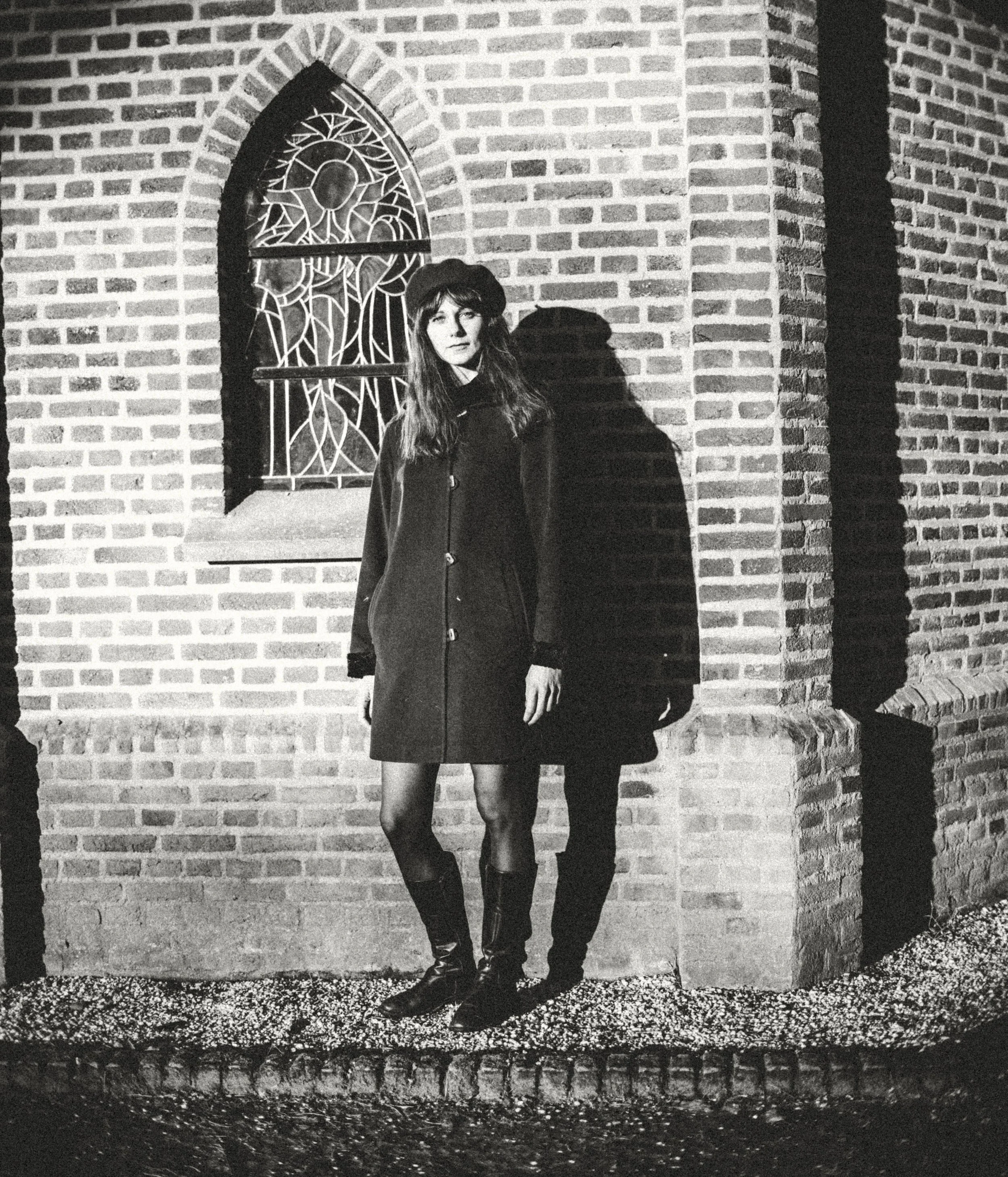 Black and white photo of a woman standing in front of a brick wall with a stained glass window. She is wearing a coat, a hat, and knee-high boots, and is looking directly at the camera. Her shadow is cast on the wall behind her.