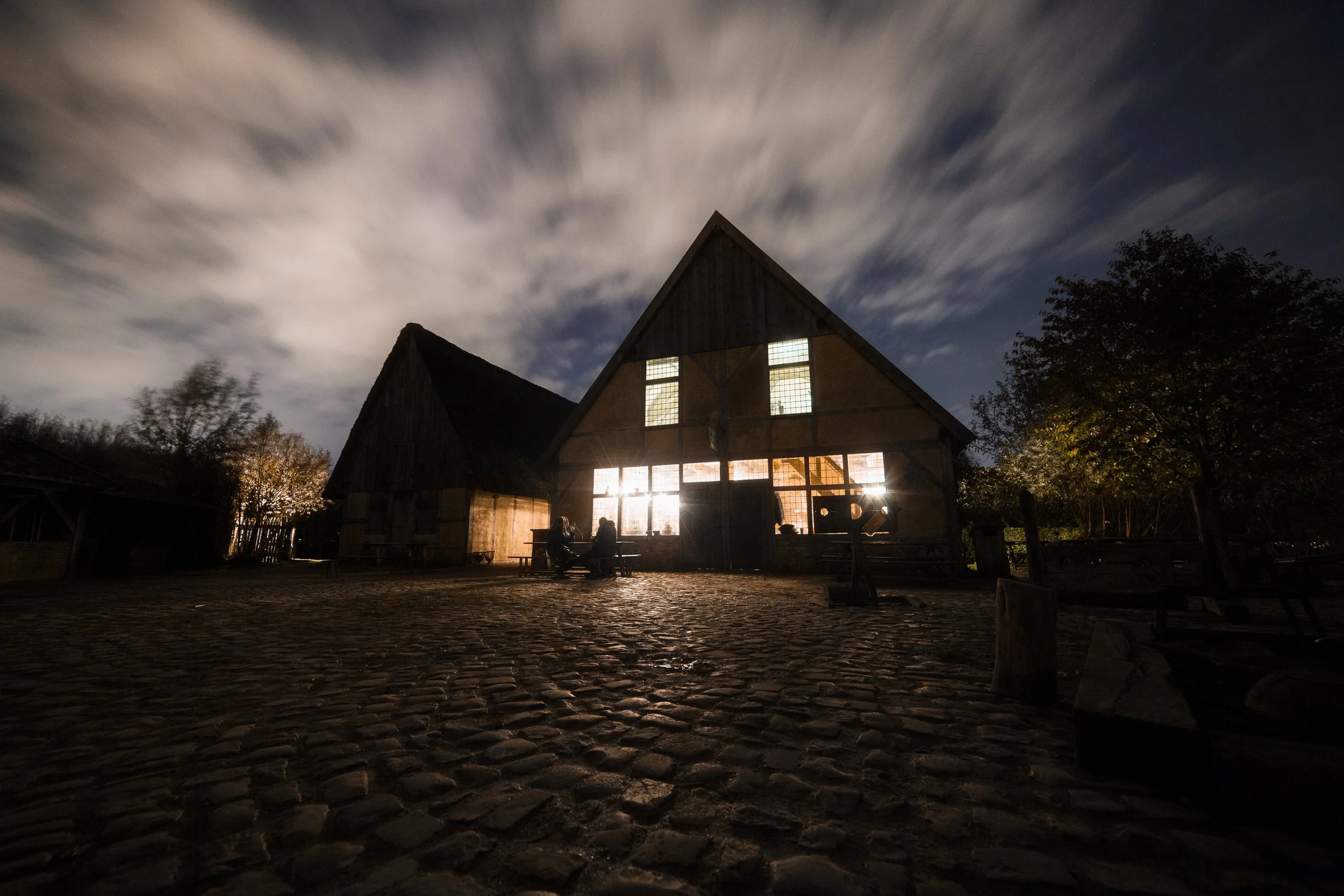 A rustic building with large windows is illuminated from the inside at night. The sky above has fast-moving clouds, creating a streaked appearance. There are trees on the right and people sitting outside near the building on cobblestone ground.