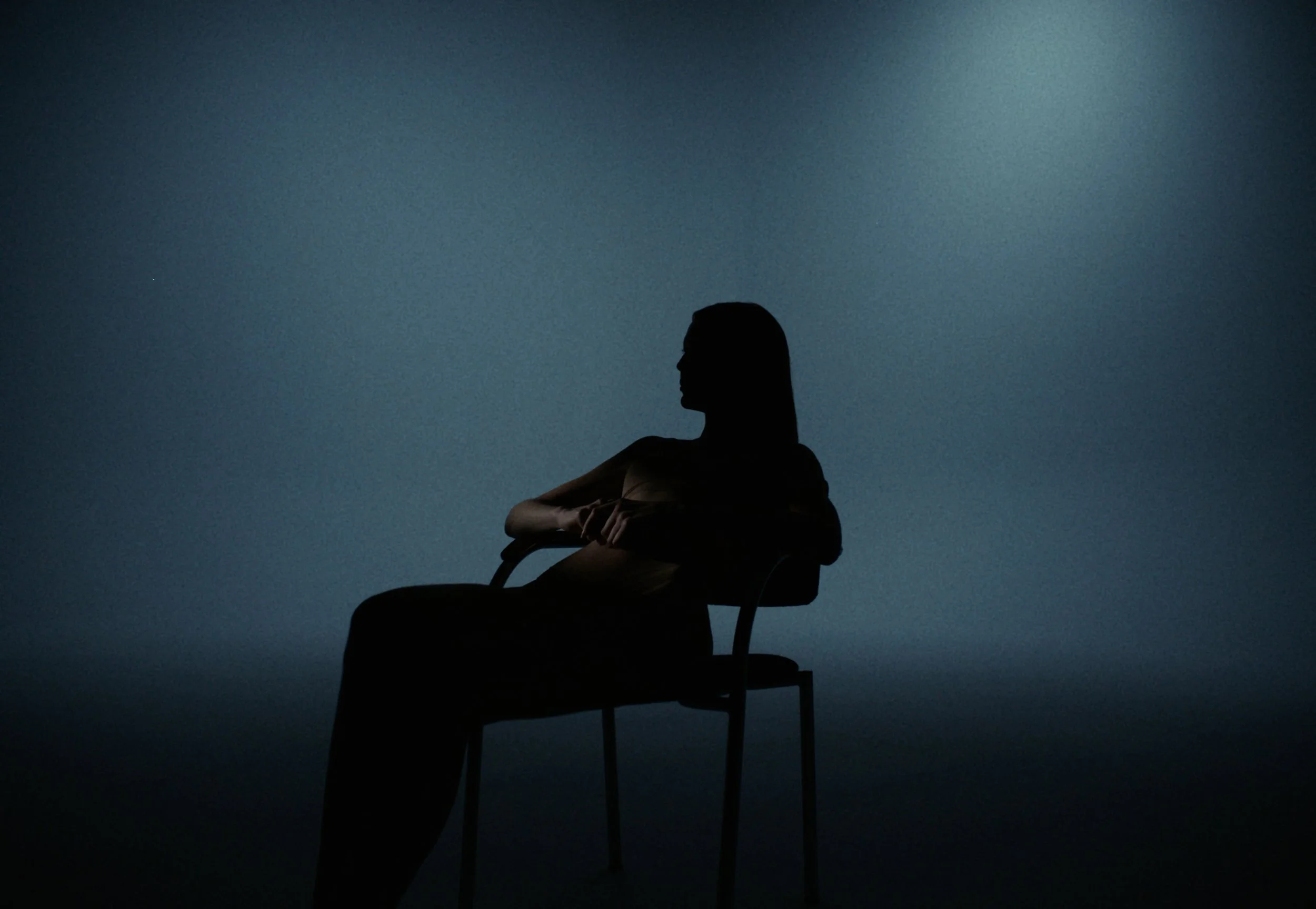 Silhouette of a person sitting on a chair against a dark background, with a gradient of blue and greenish hues.