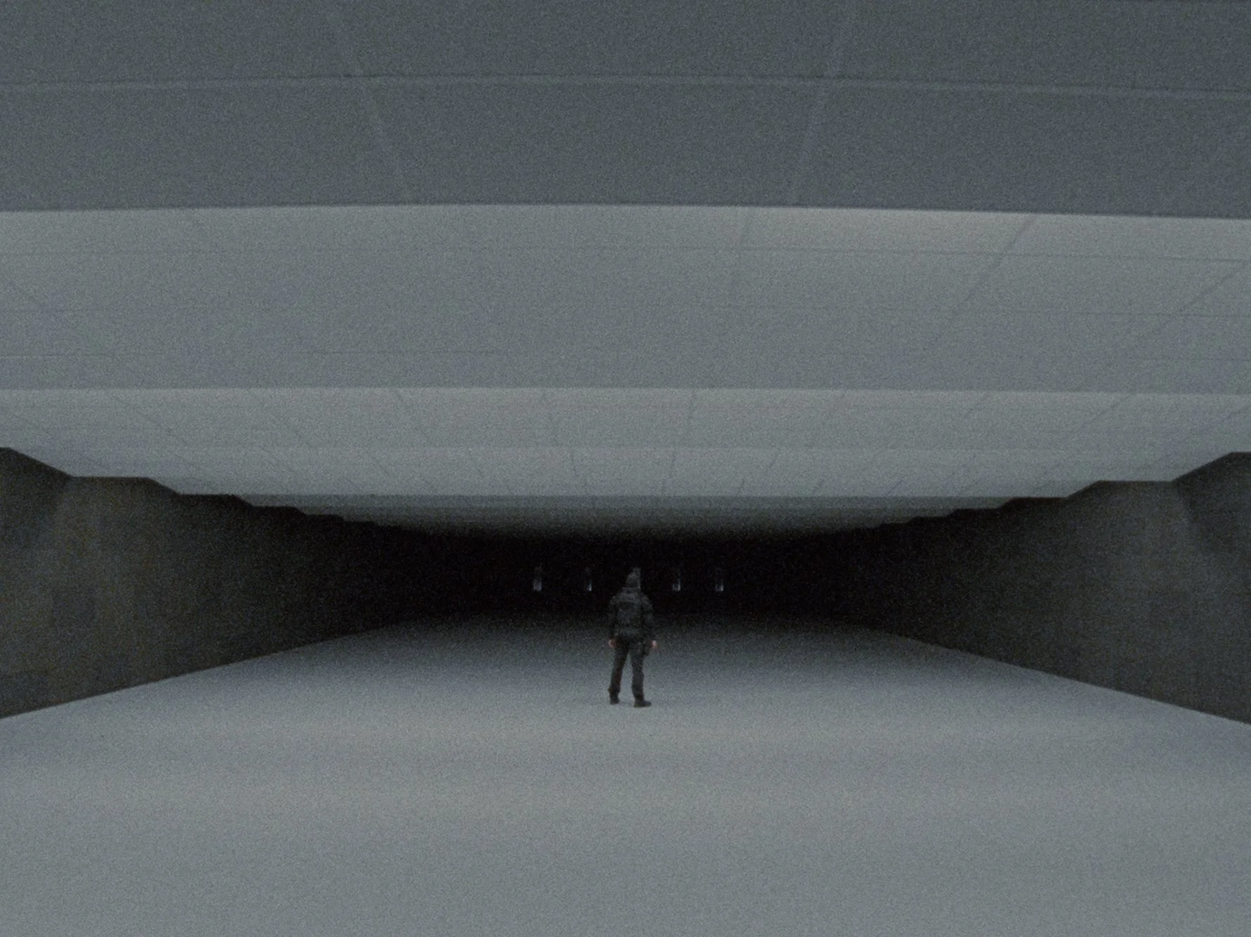 A person standing in the center of an empty, dark, indoor space with a high ceiling, surrounded by a curved wall or screen, with soft lighting illuminating the scene.