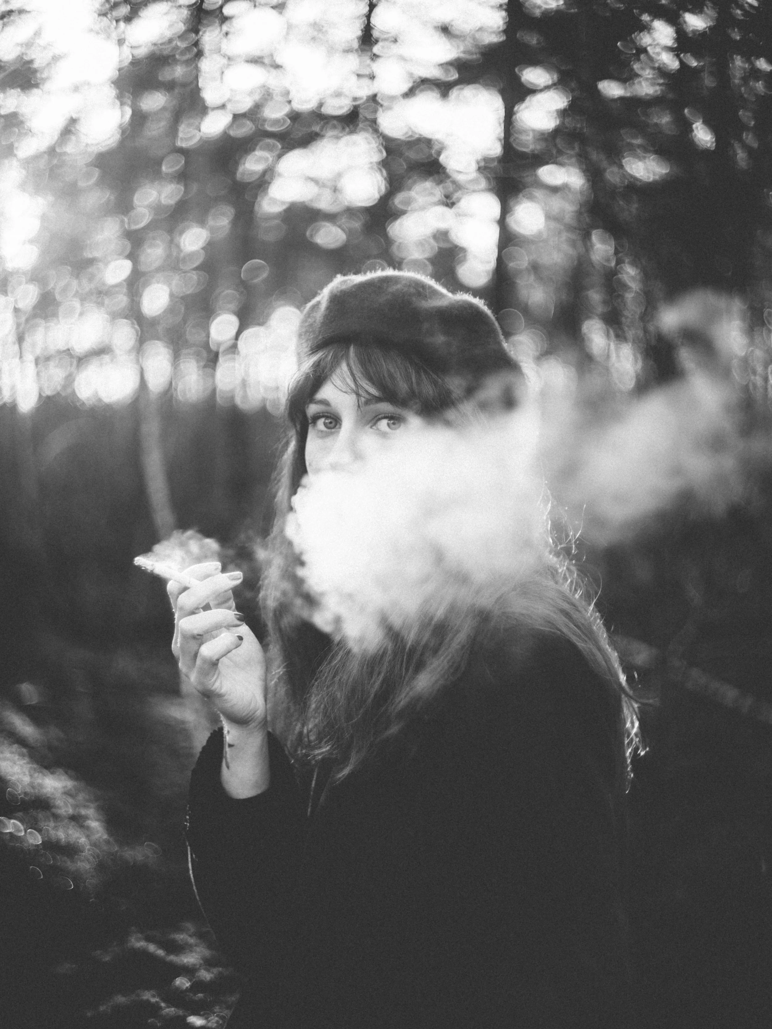 A woman with dark hair and light eyes holding a cigarette, wearing a beret and a coat with fur trim, outdoors with a blurred background of trees and bokeh lights.