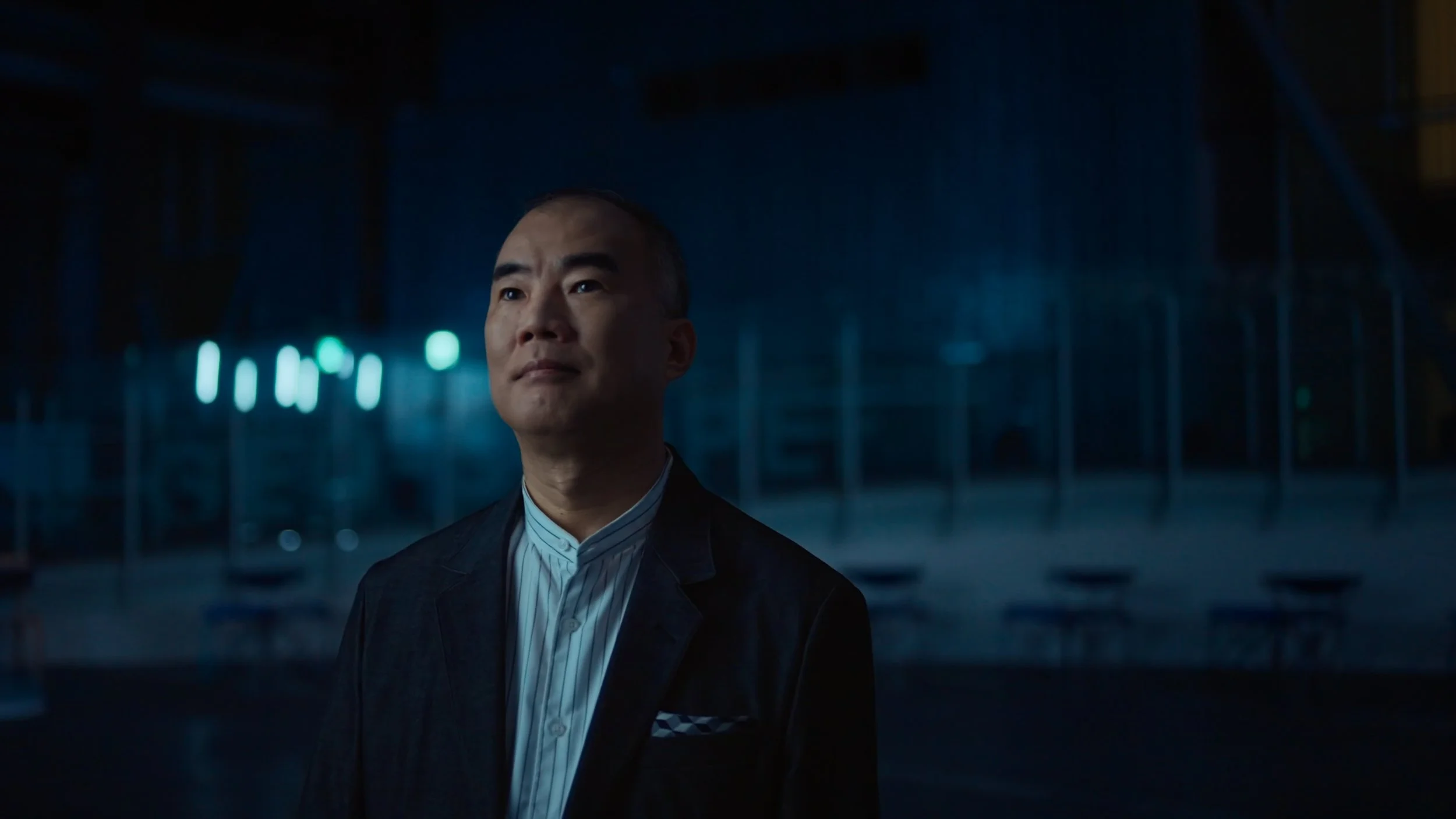A man in a suit standing in a dark, industrial-looking space with teal lights in the background.