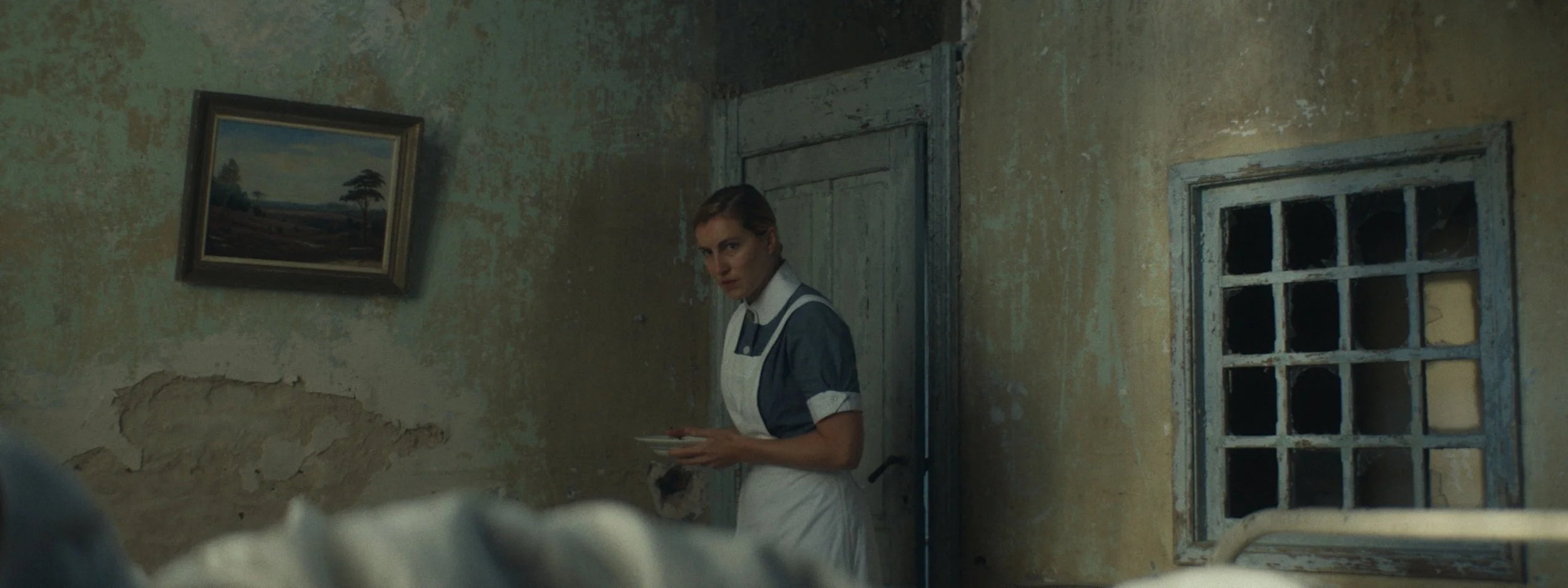 A woman in a vintage-style maid uniform holding a plate, standing in a dilapidated room with peeling walls, an old window, and a landscape painting on the wall.