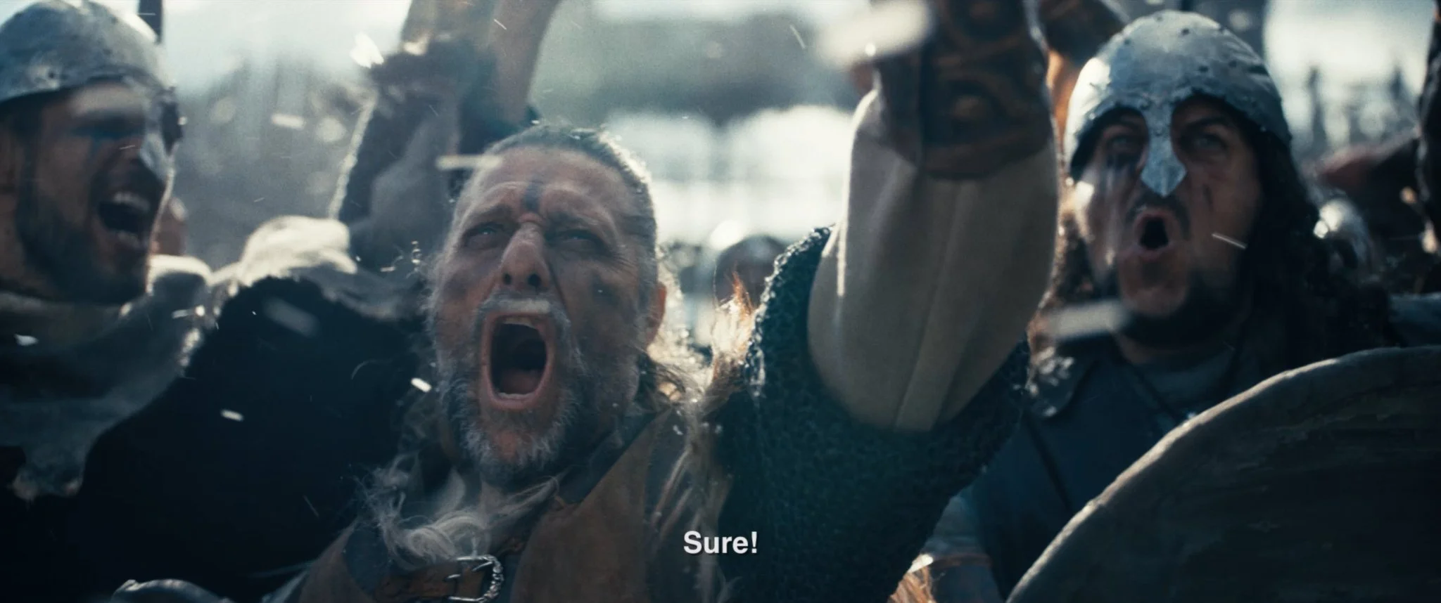 A group of people, some with face paint, appear to be in a chaotic or protest scene. The person in the center has long hair and a beard, with an intense expression, mouth open as if shouting. Others around are also shouting or yelling, and some are w