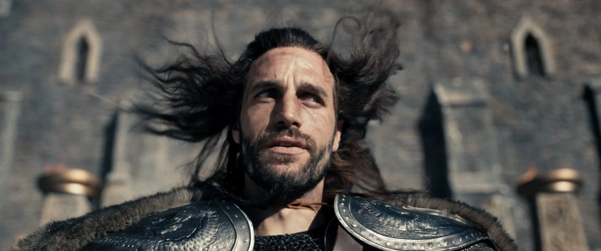 A man with long hair and beard lying on the ground, wearing medieval armor and a fur cloak, with a castle wall in the background.