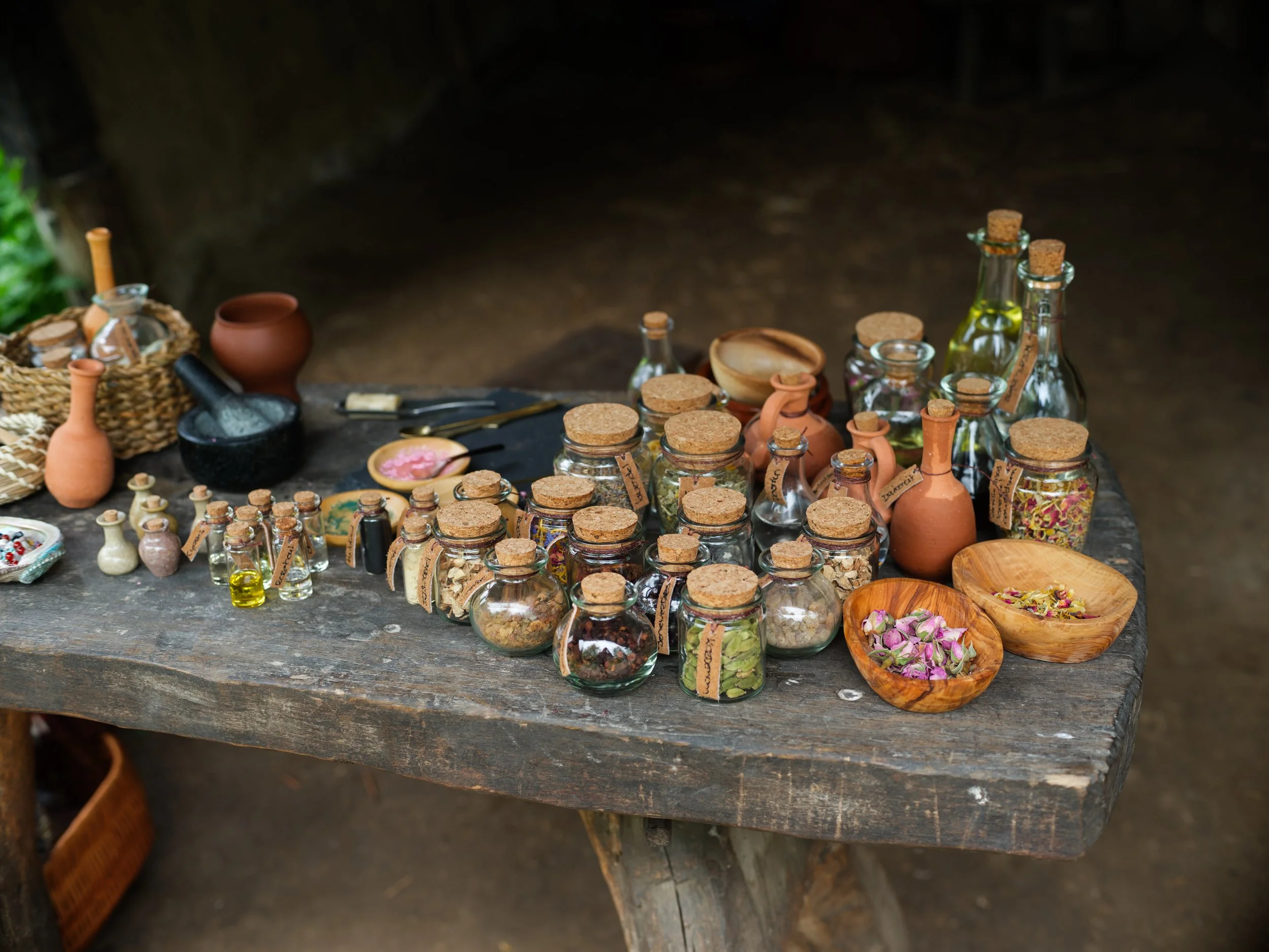 A rustic wooden table displays various small glass jars with cork stoppers filled with colorful herbs, spices, and dried flowers, alongside small clay pots, a small spoon, and a woven basket.