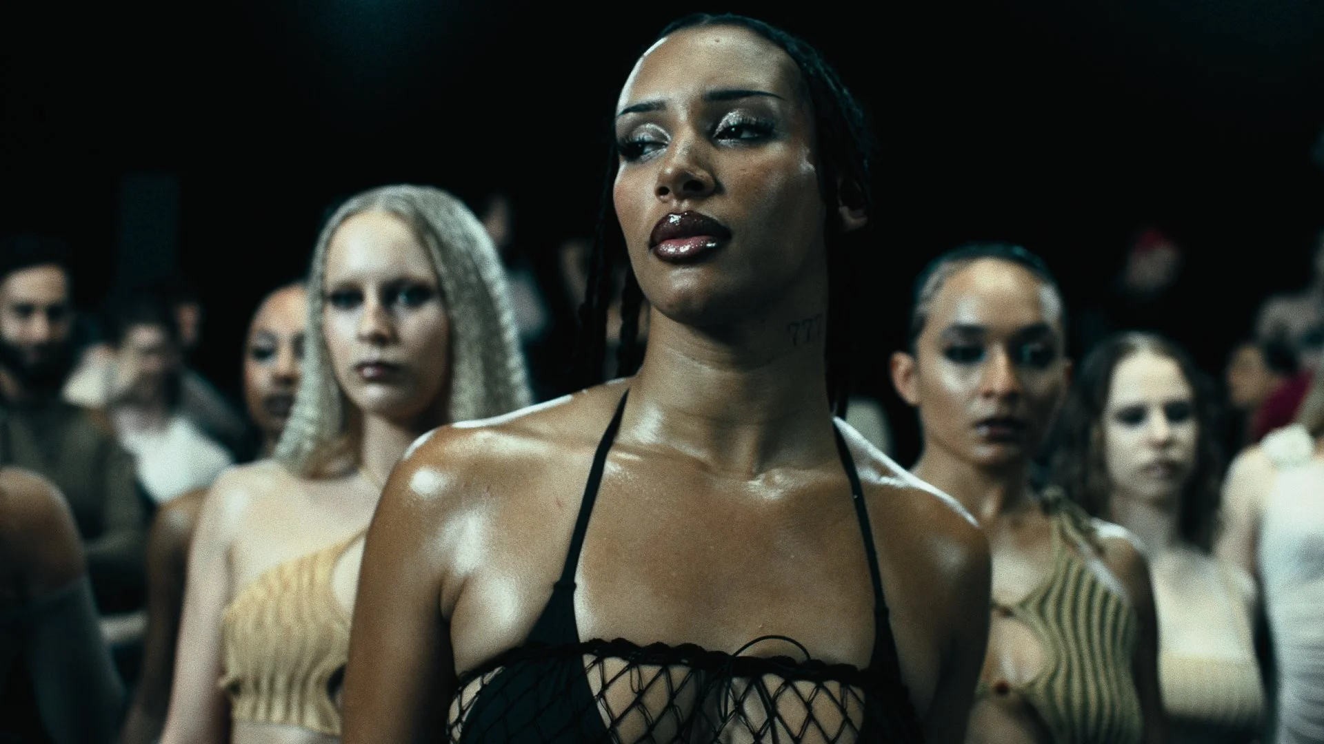 A group of women standing together, with one woman in focus in front, wearing a black strappy top, with braided hair and makeup, surrounded by others in a dimly lit setting.