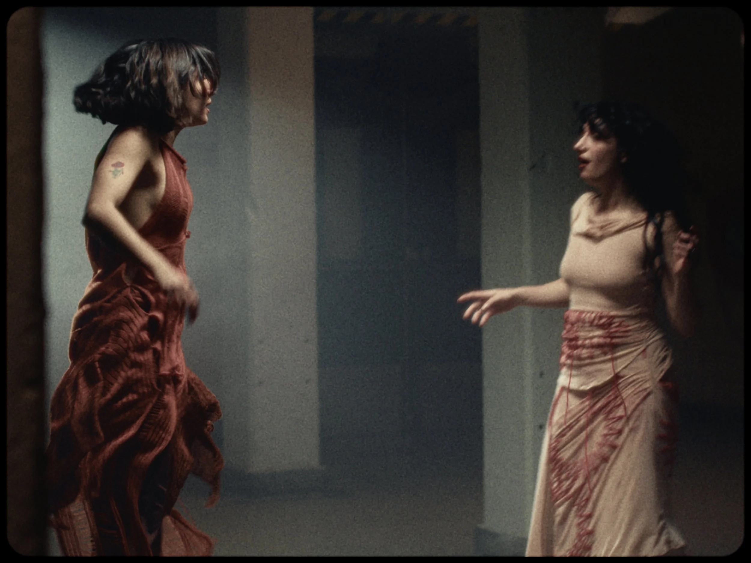 Two women are facing each other in a dimly lit room. The woman on the left has short, wavy brown hair, wears a red sleeveless top, and has a small tattoo on her left arm. The woman on the right has long, dark curly hair, and wears a light-colored top
