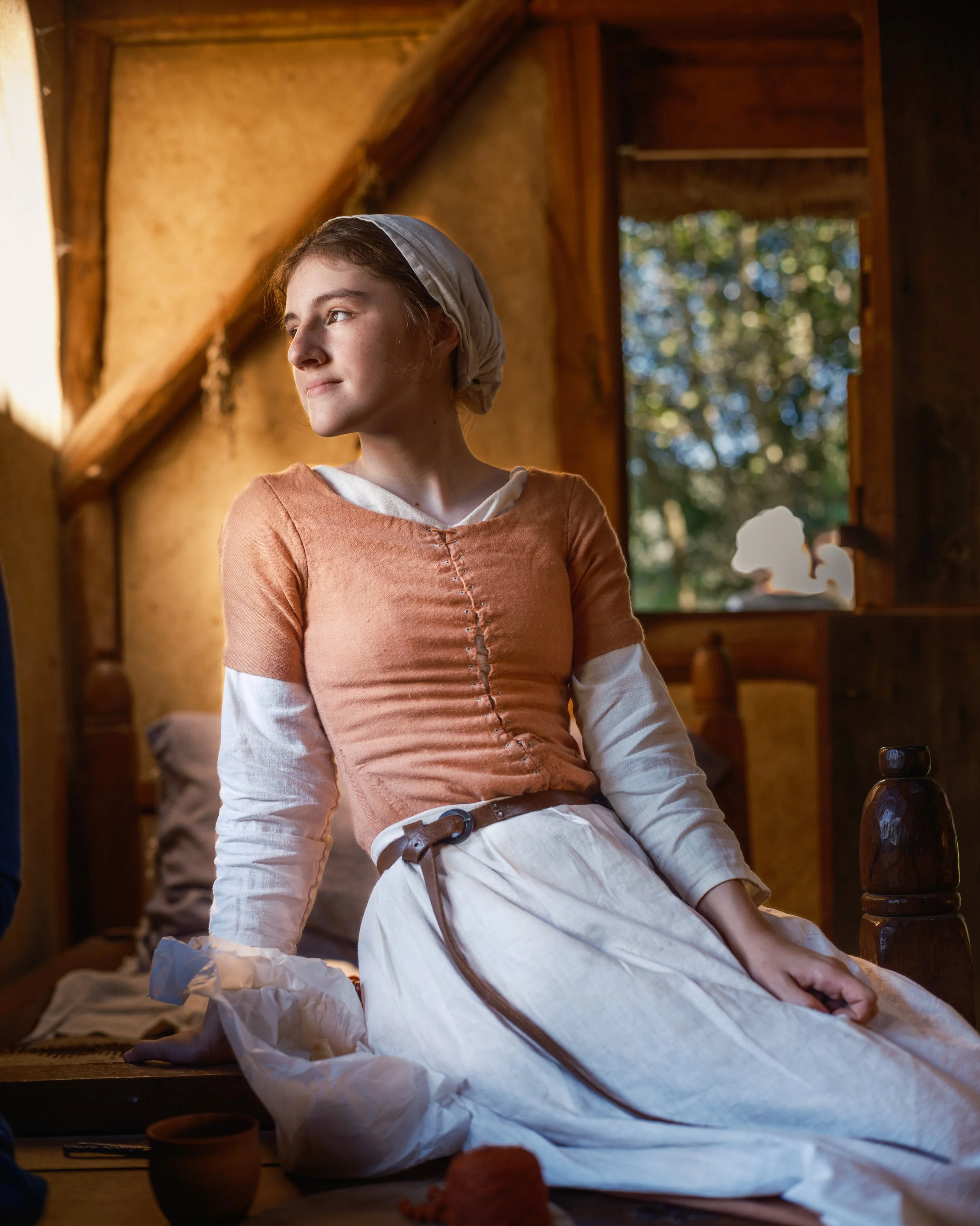 A woman dressed in historical clothing, sitting in a rustic wooden room with a large window showing trees outside, with crumpled paper and pottery on the floor.