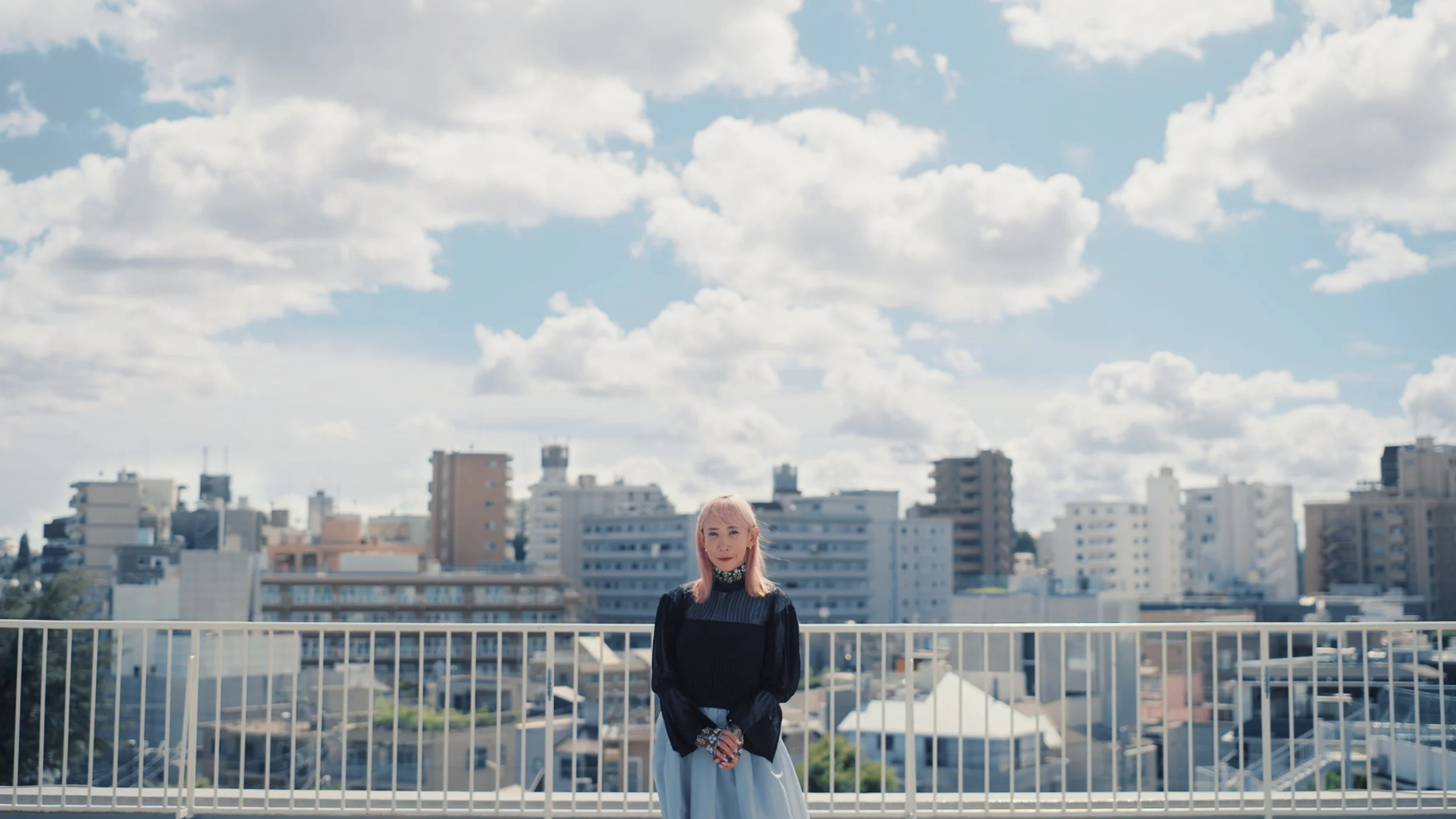 A woman with pink hair standing on a rooftop balcony in an urban area under a cloudy sky.
