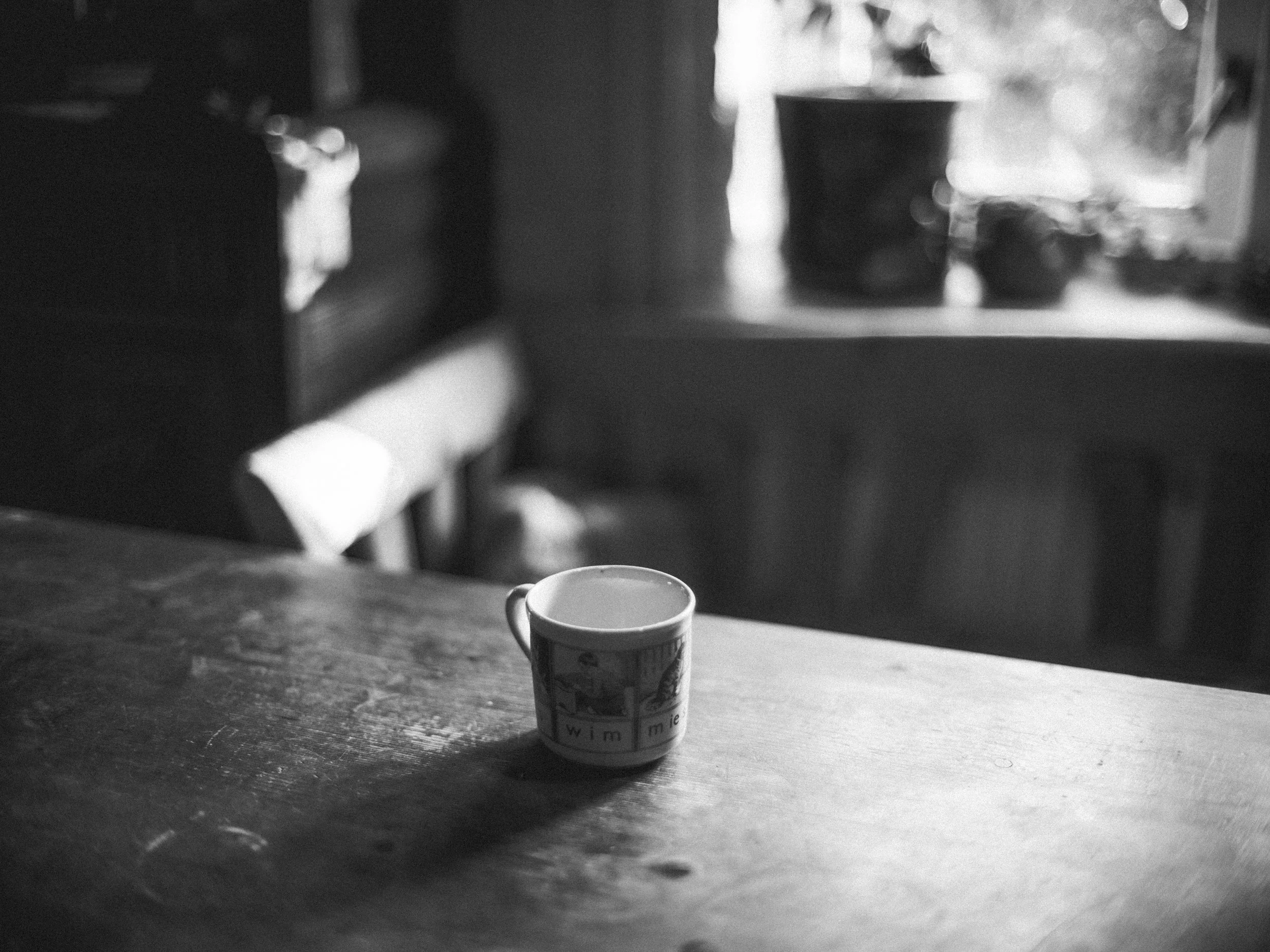 A black-and-white photo of a coffee cup on a wooden table with a blurred background, including a second cup and a window.