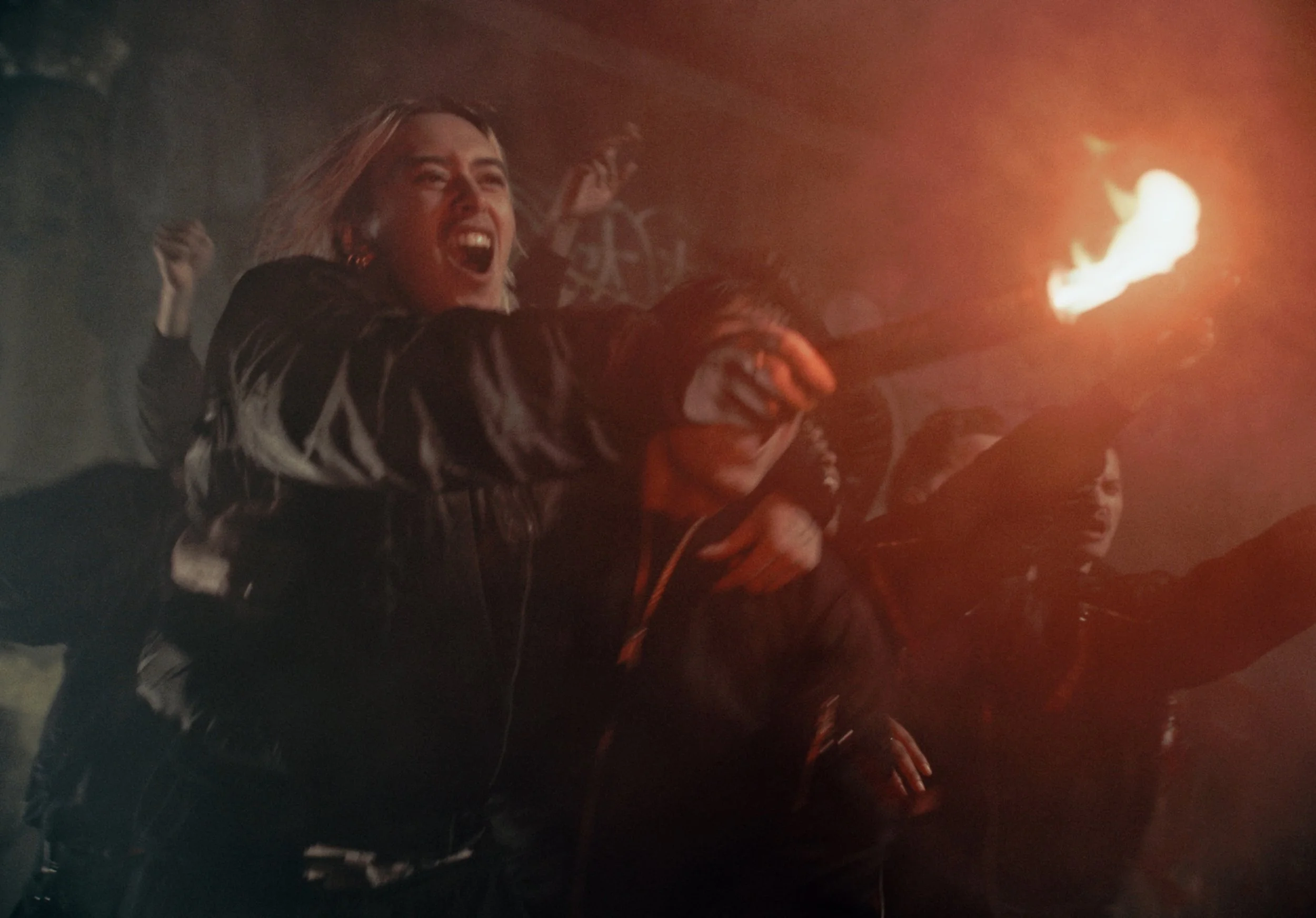 A group of people, mostly women, joyfully holding torches with flames at night, with one woman in the foreground laughing and raising her hand.