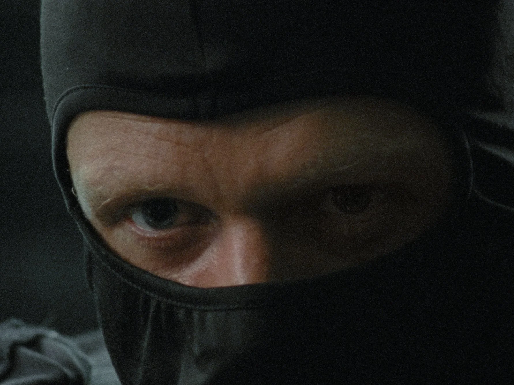 Close-up of a person's face wearing a black mask and black cap, with one eye and part of the forehead visible.