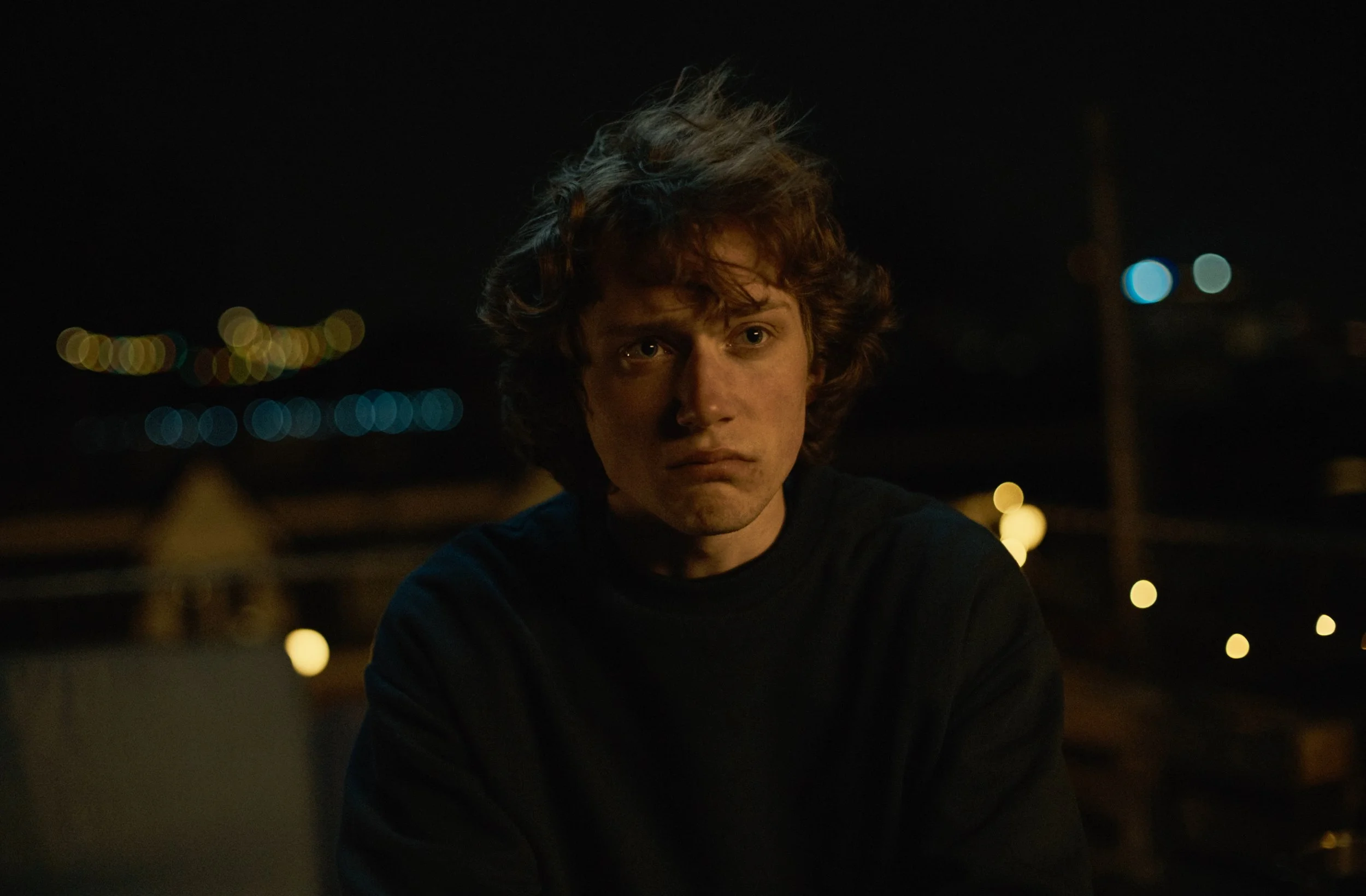 A young man with curly hair is looking into the camera at night, with blurred city lights in the background.