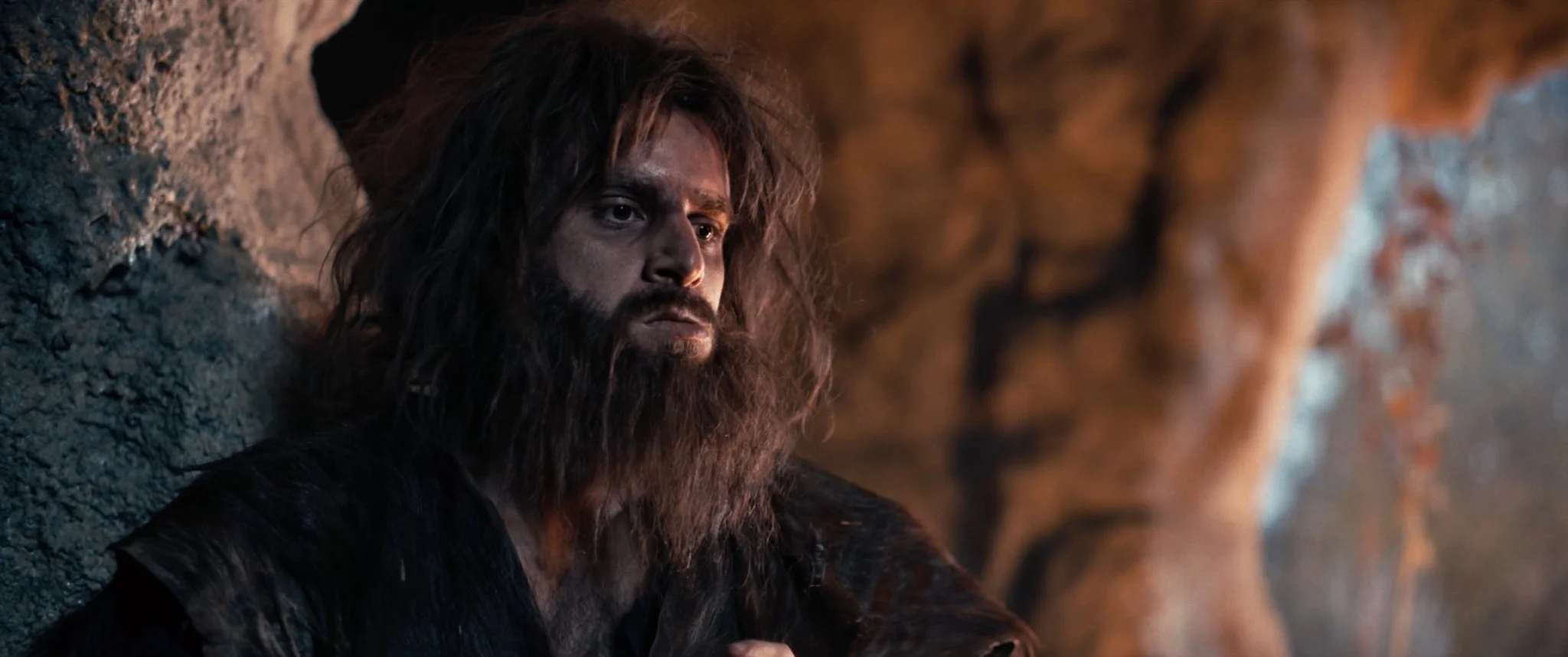 A man with long hair and a beard resting against a rock wall, looking pensive with a blurred rocky background.