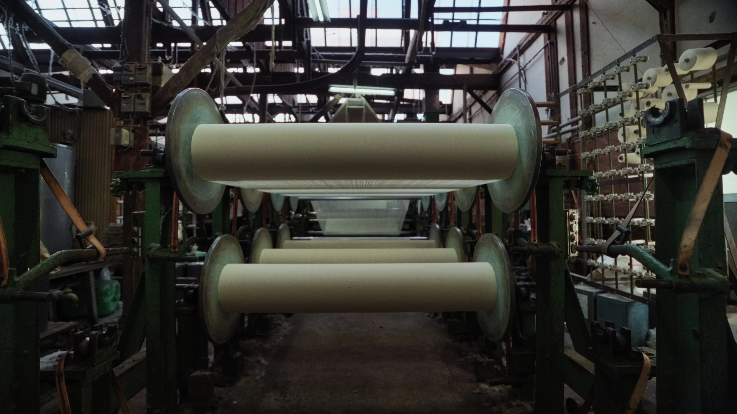 Industrial machinery with multiple large beige rollers in a factory setting, with a dark interior and metal framework above.