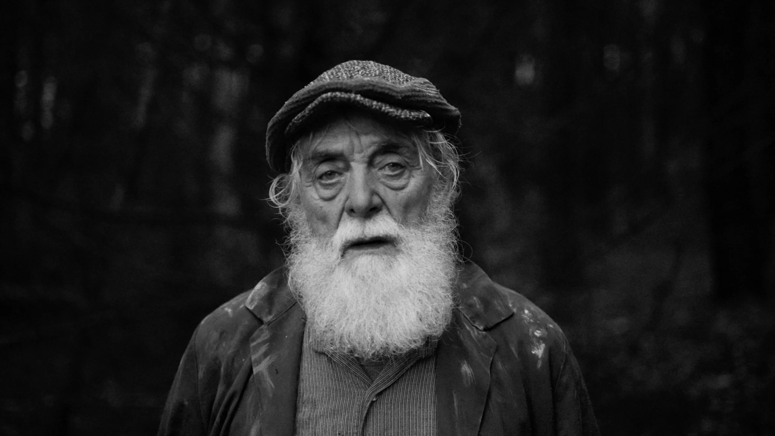 Black and white photo of an elderly man with a long white beard, wearing a hat and a jacket, standing outdoors in a forested area.